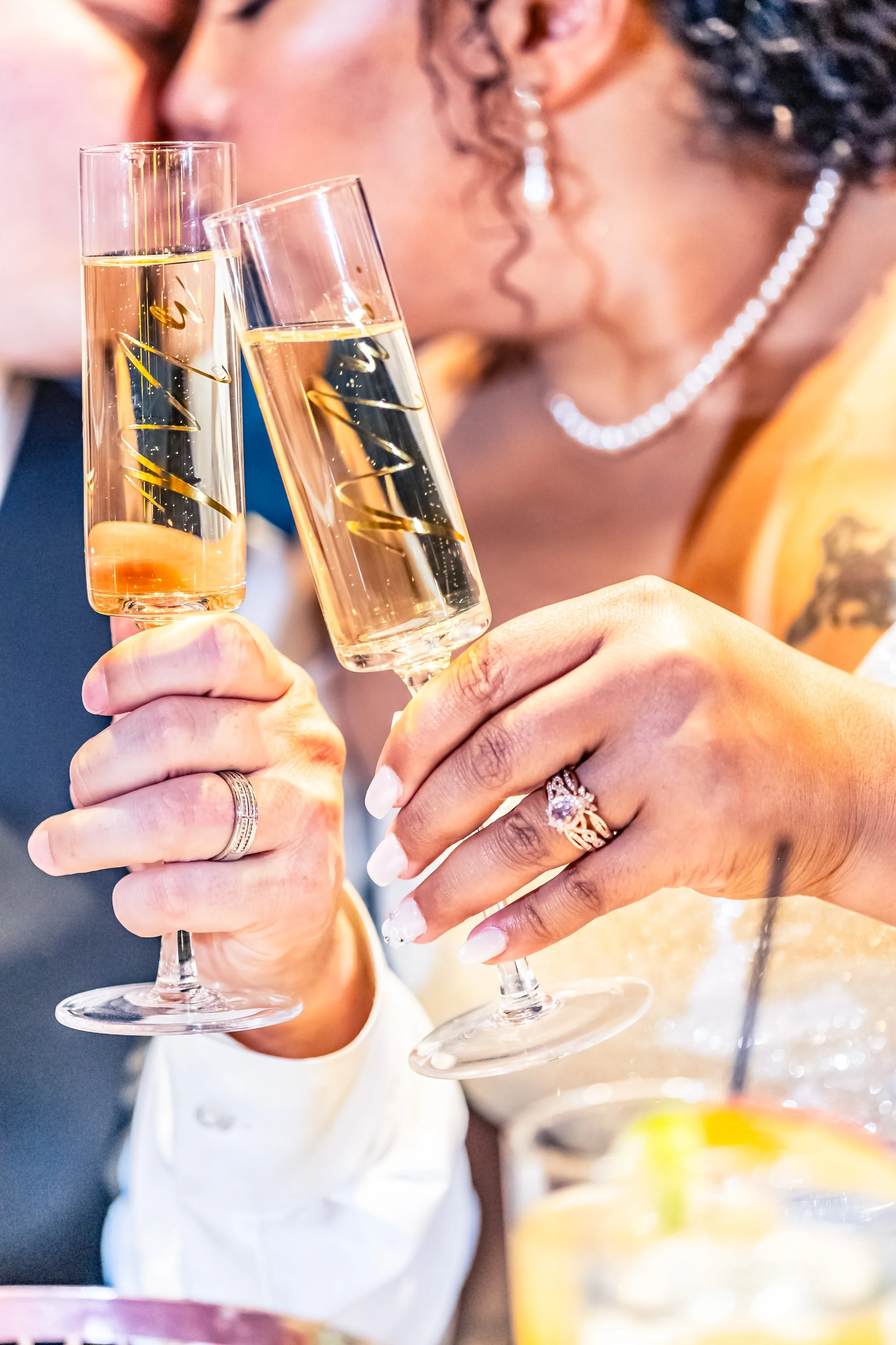 Close-up of a couple toasting with champagne glasses at a celebration or wedding, with a woman's hand showing jewelry, including a wedding ring and engagement ring.