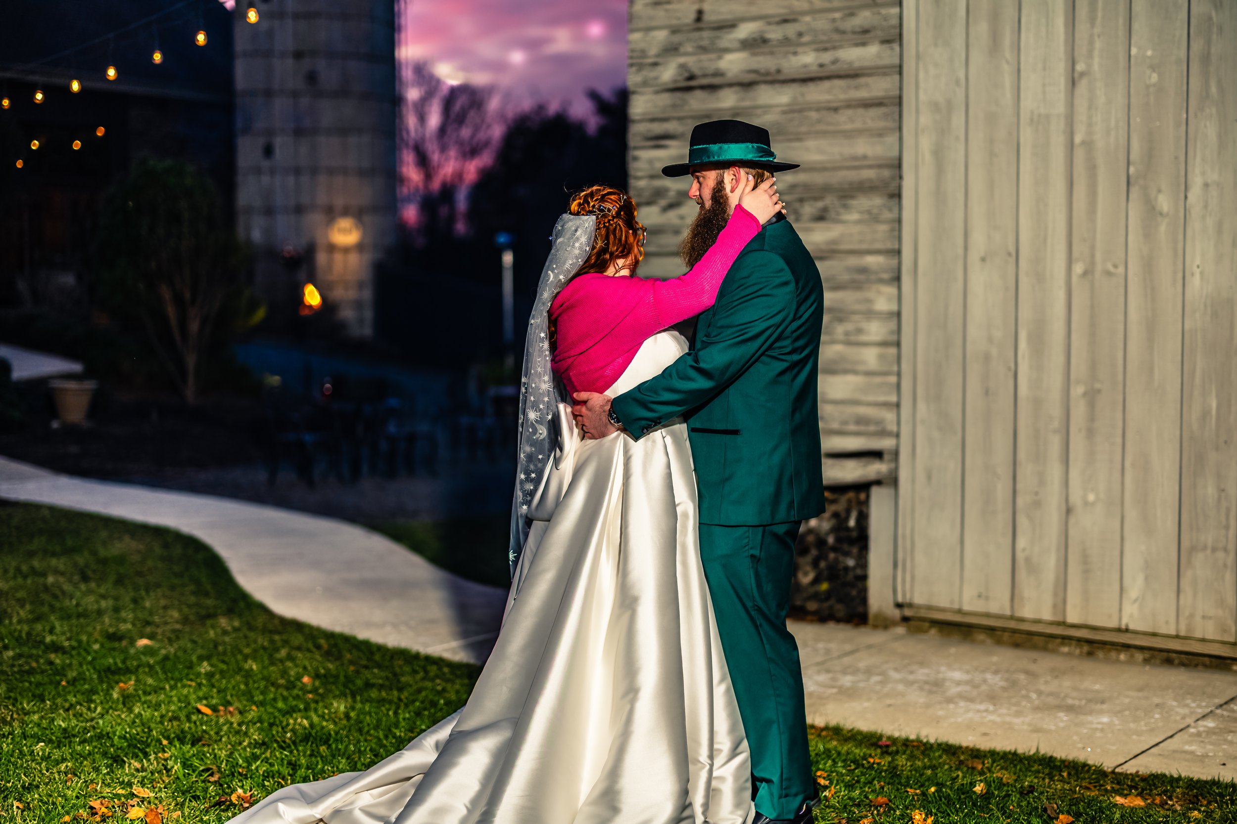 A bride and groom sharing a dance outside during dusk, with the bride wearing a satin wedding gown and a veil, and the groom in a teal suit and black hat. The bride has her arms around the groom's neck, and the groom is holding her waist.