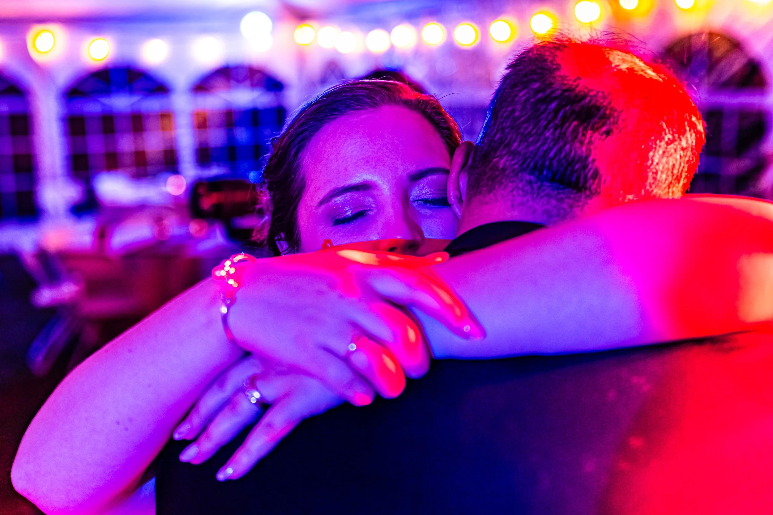 A woman and a man hugging closely at a party or dance event, with colorful purple and red lighting in the background.