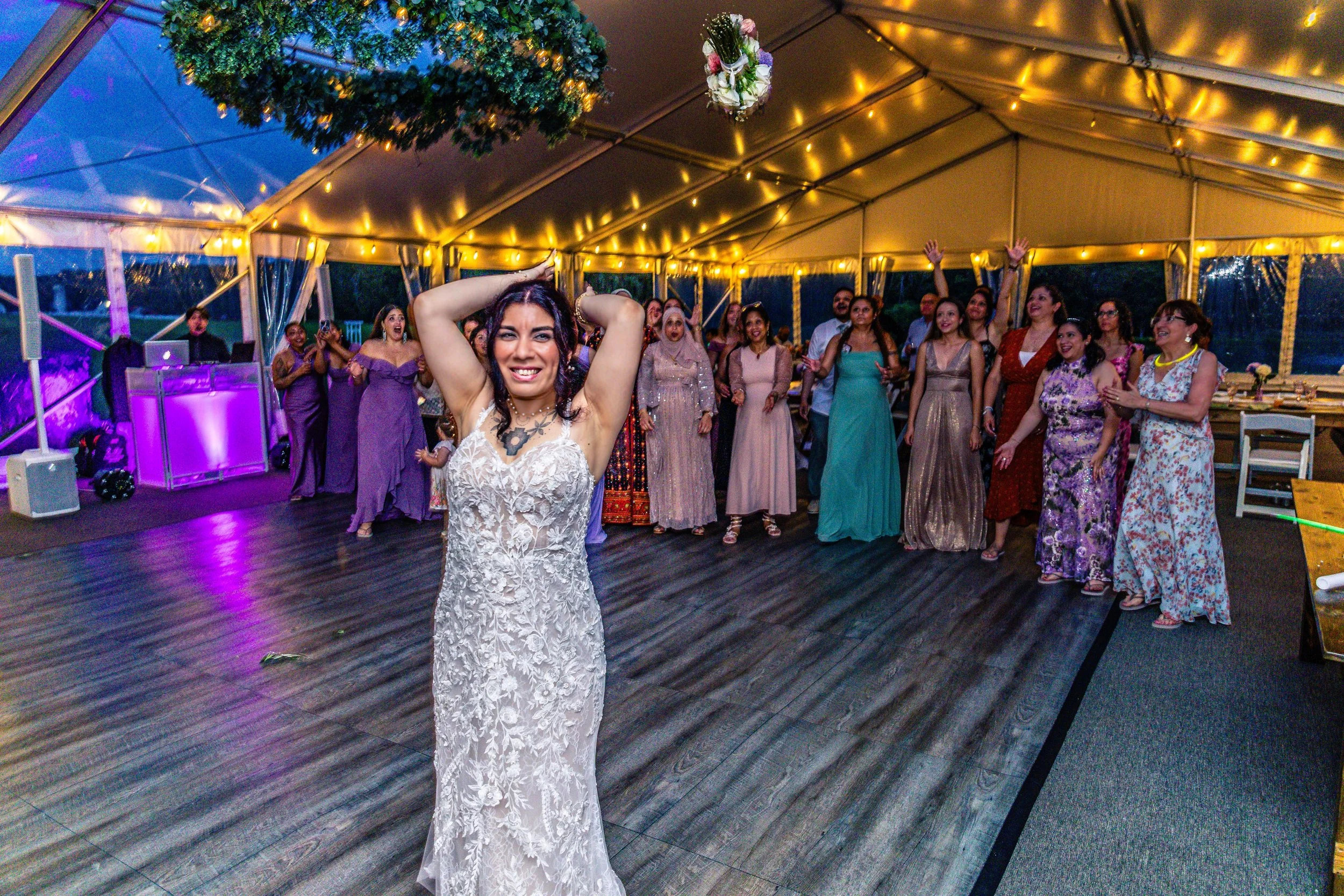 A woman in a white lace dress stands in the center of a dance floor, smiling with arms raised behind her head, at a celebration with people dancing and enjoying themselves under a decorated tent with string lights.