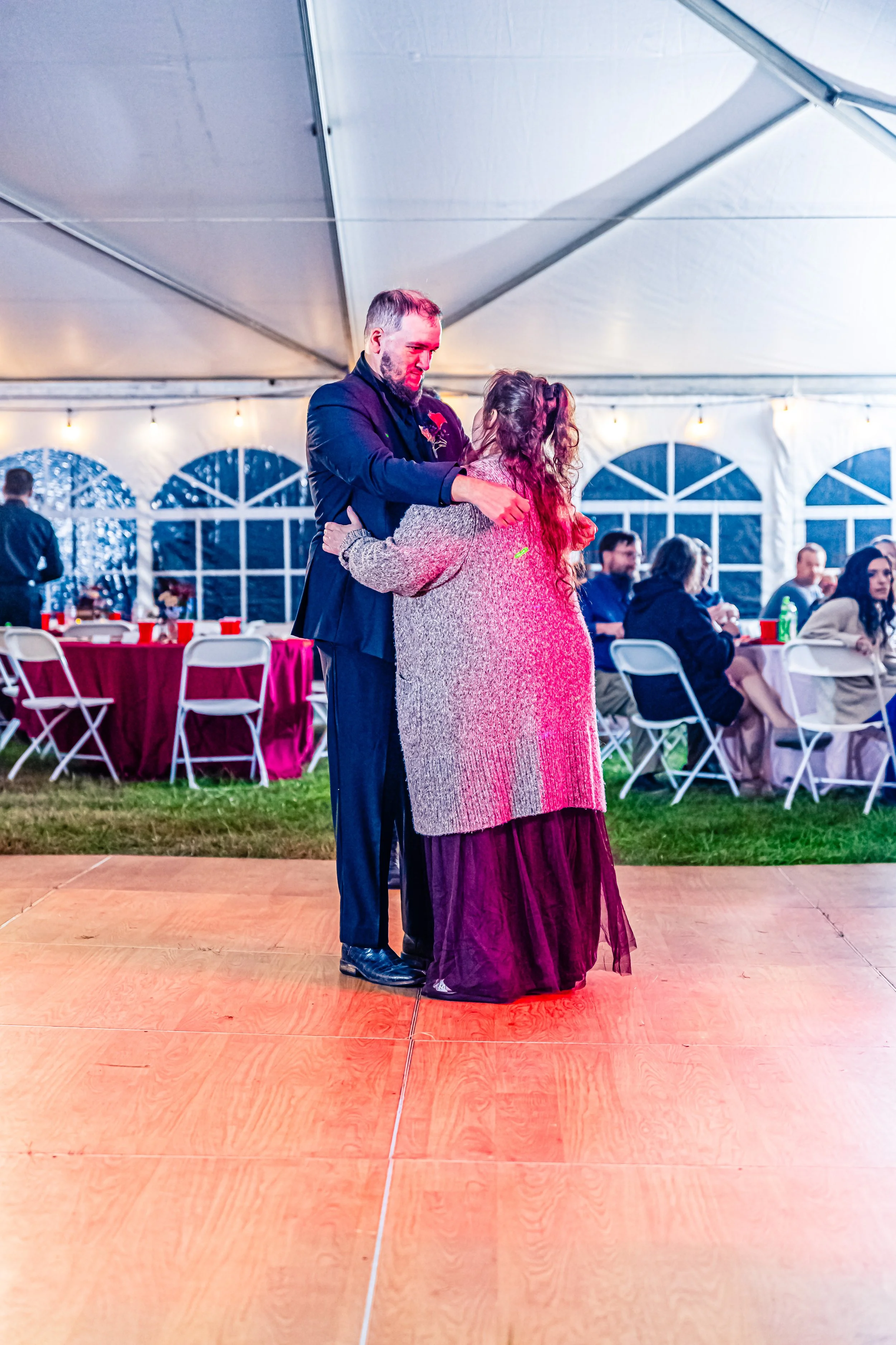 A couple dancing together at a wedding reception under a large white event tent, with tables and guests seated in the background.