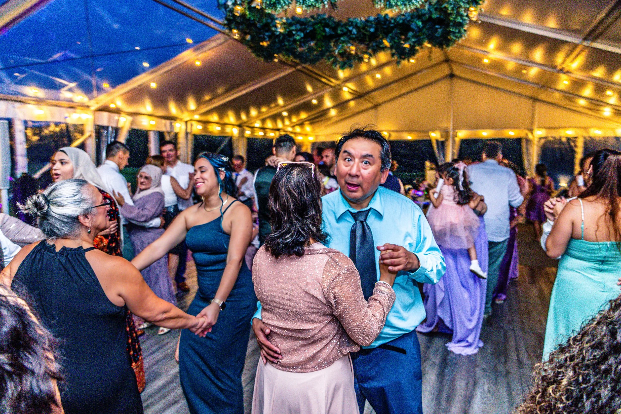 People dancing and socializing under a tent with string lights at a festive event.