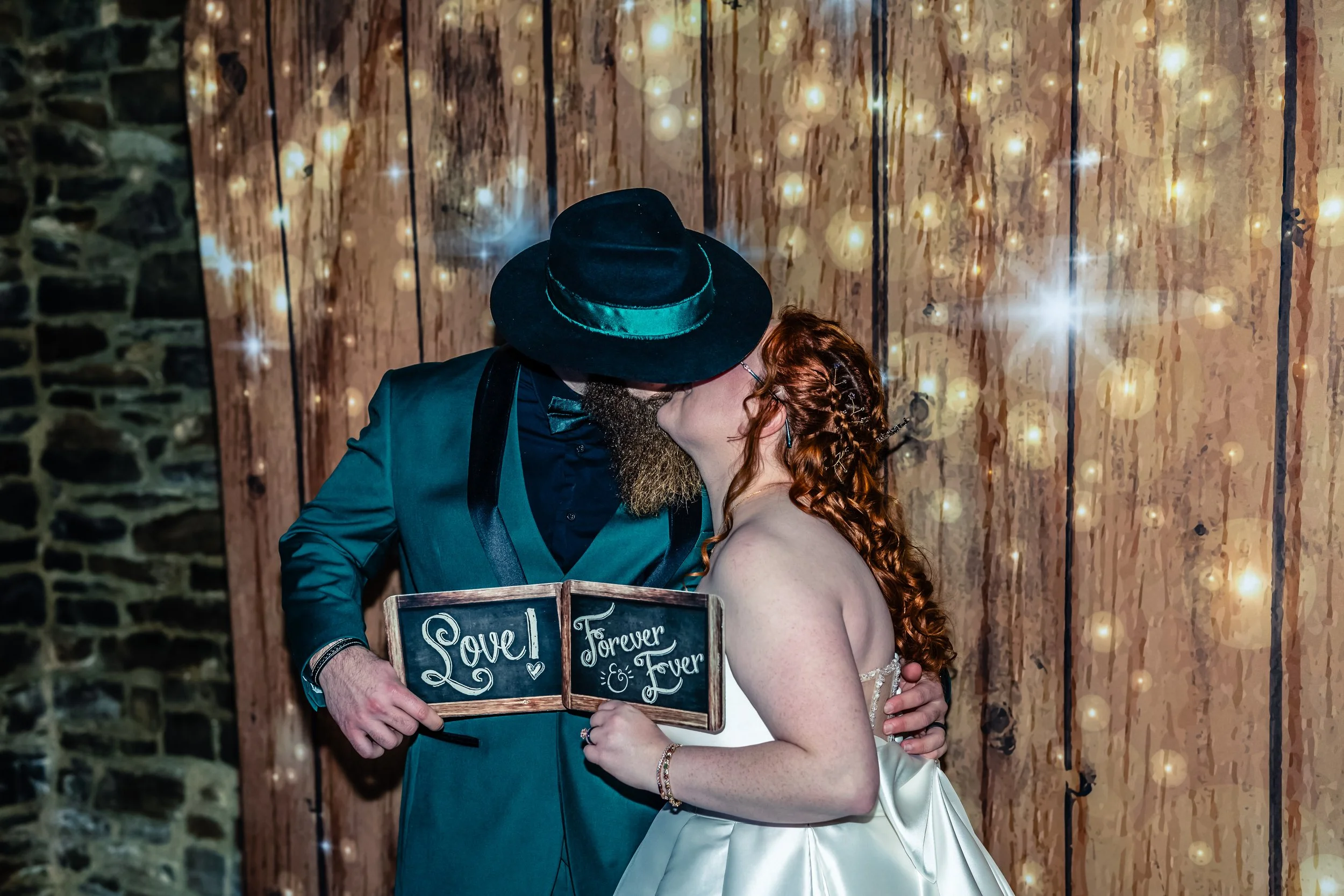 A couple kissing at a wedding reception, holding a small chalkboard sign that reads 'Love Forever & Fever.' The man has a beard and is wearing a dark suit, tie, and a wide-brimmed black hat. The woman has long curly red hair and is wearing a white sa