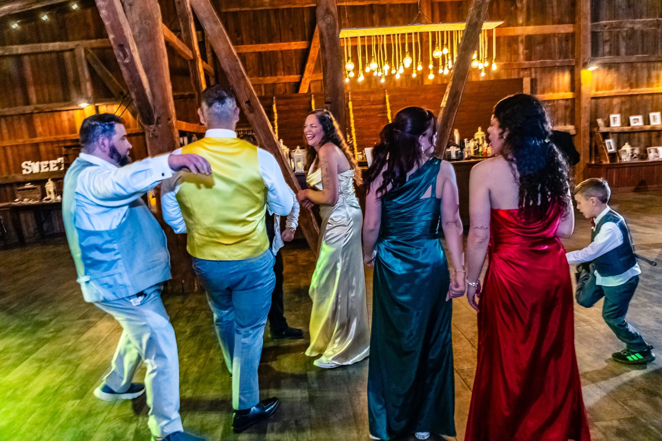 People dancing in a barn wedding reception, with four women in colorful dresses and two men in suits, and a young boy in a vest and pants dancing nearby.
