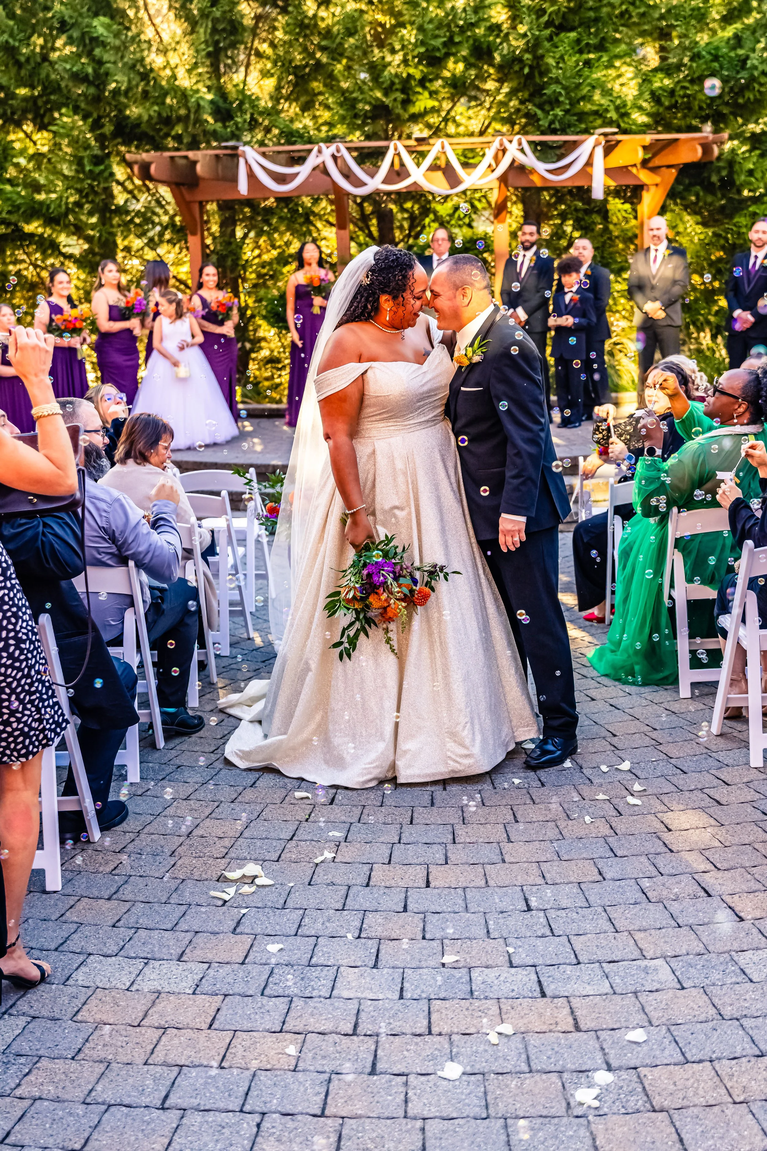 A wedding ceremony outdoors with a bride and groom touching foreheads, surrounded by guests, with wedding party members in the background, trees and a decorated wooden structure, and bubbles floating in the air.