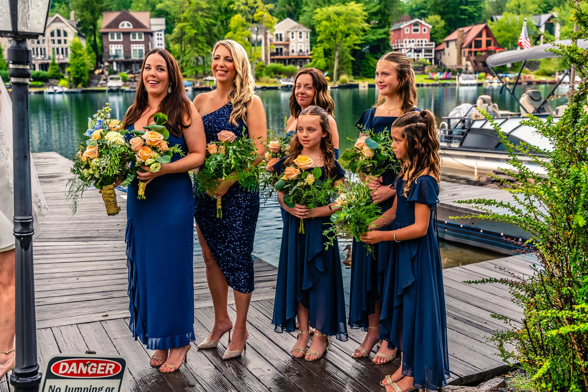 A group of six women and girls dressed in blue dresses holding bouquets, standing on a wooden dock near a lake with boats and lakeside houses in the background, likely during a wedding or special occasion.