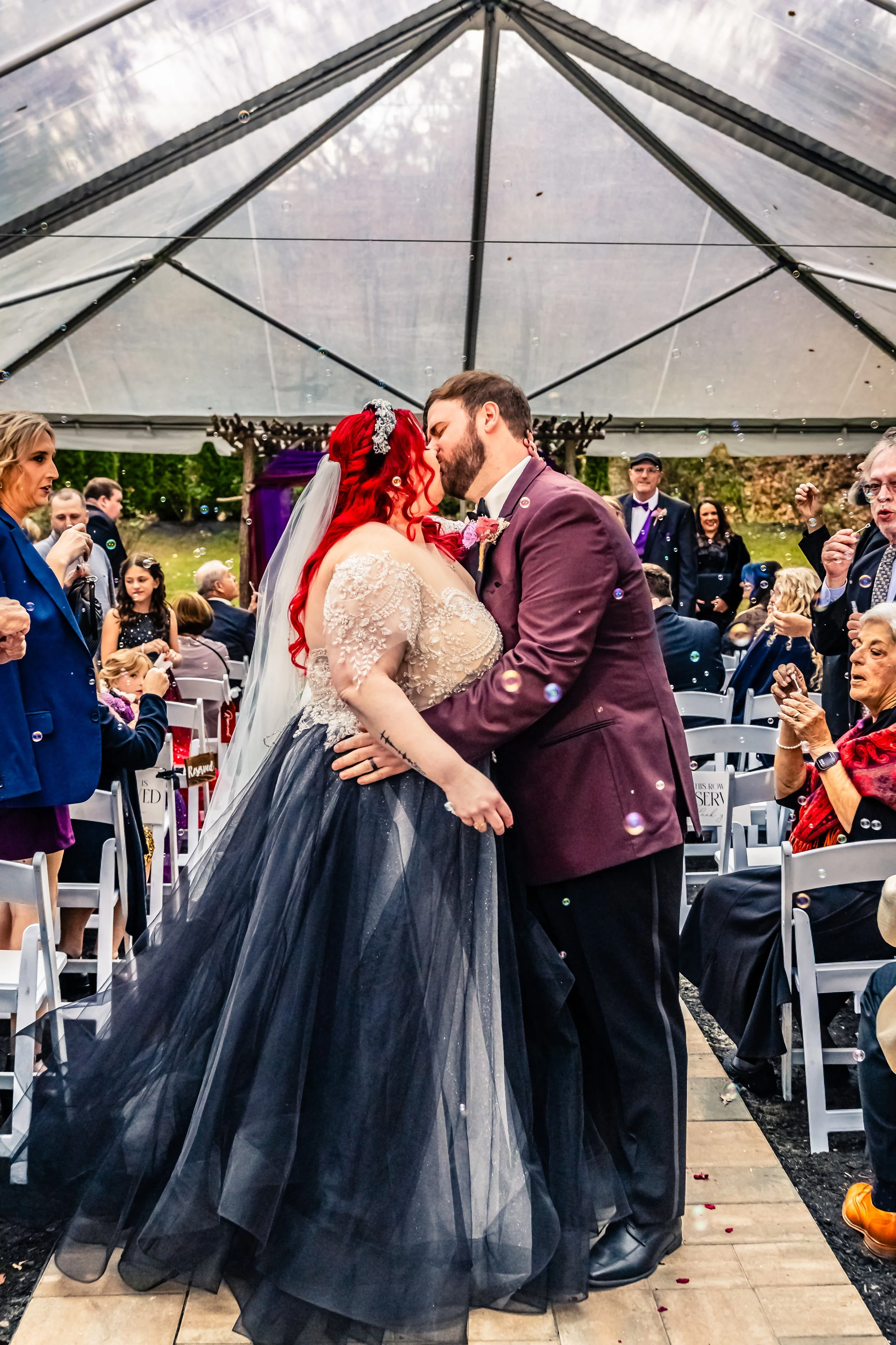 Couple sharing a kiss at their wedding ceremony under a clear tent, with seated guests around them.
