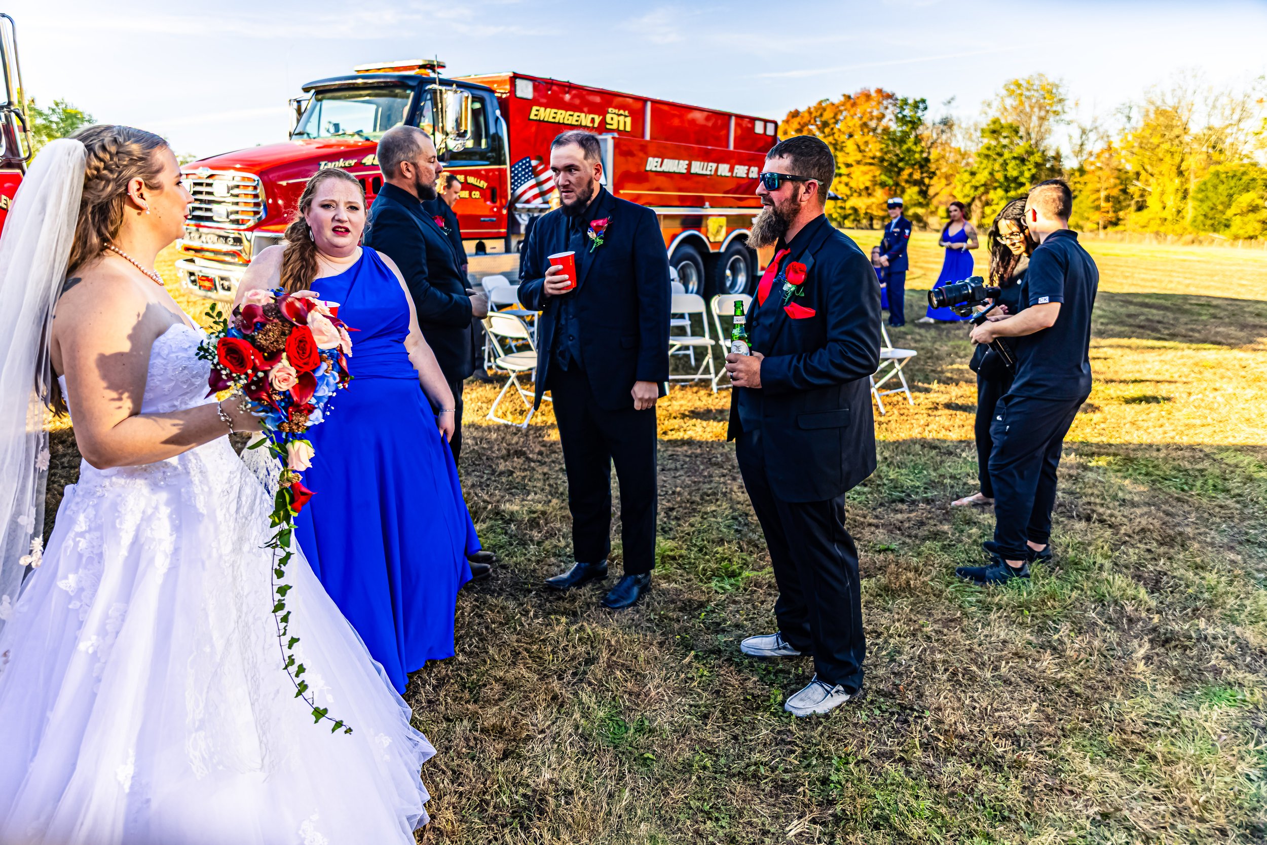 Bride in a white wedding dress holding a bouquet, talking to two women and two men in formal attire at an outdoor wedding, with a fire truck in the background.