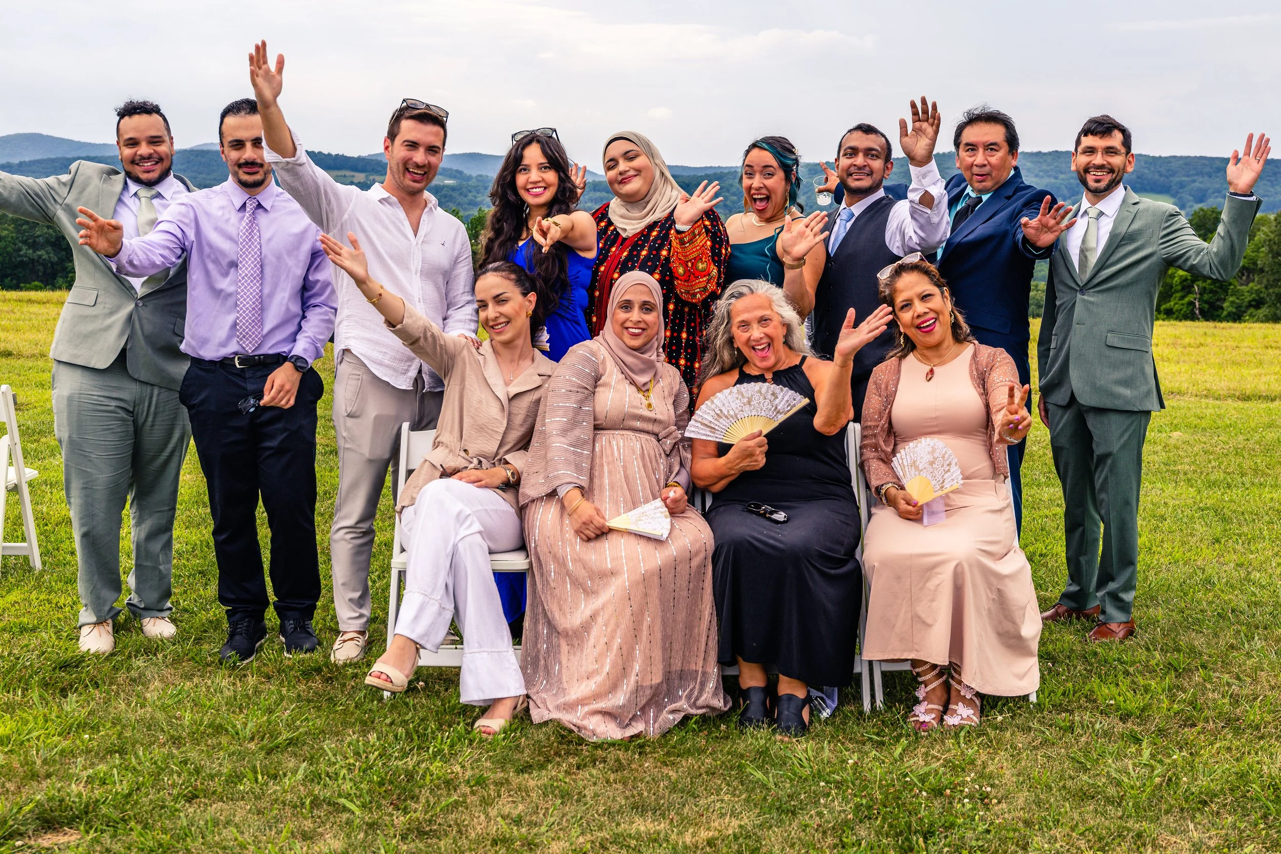 A diverse group of people dressed in formal attire, celebrating outdoors on a grassy field with mountains in the background, smiling and waving.