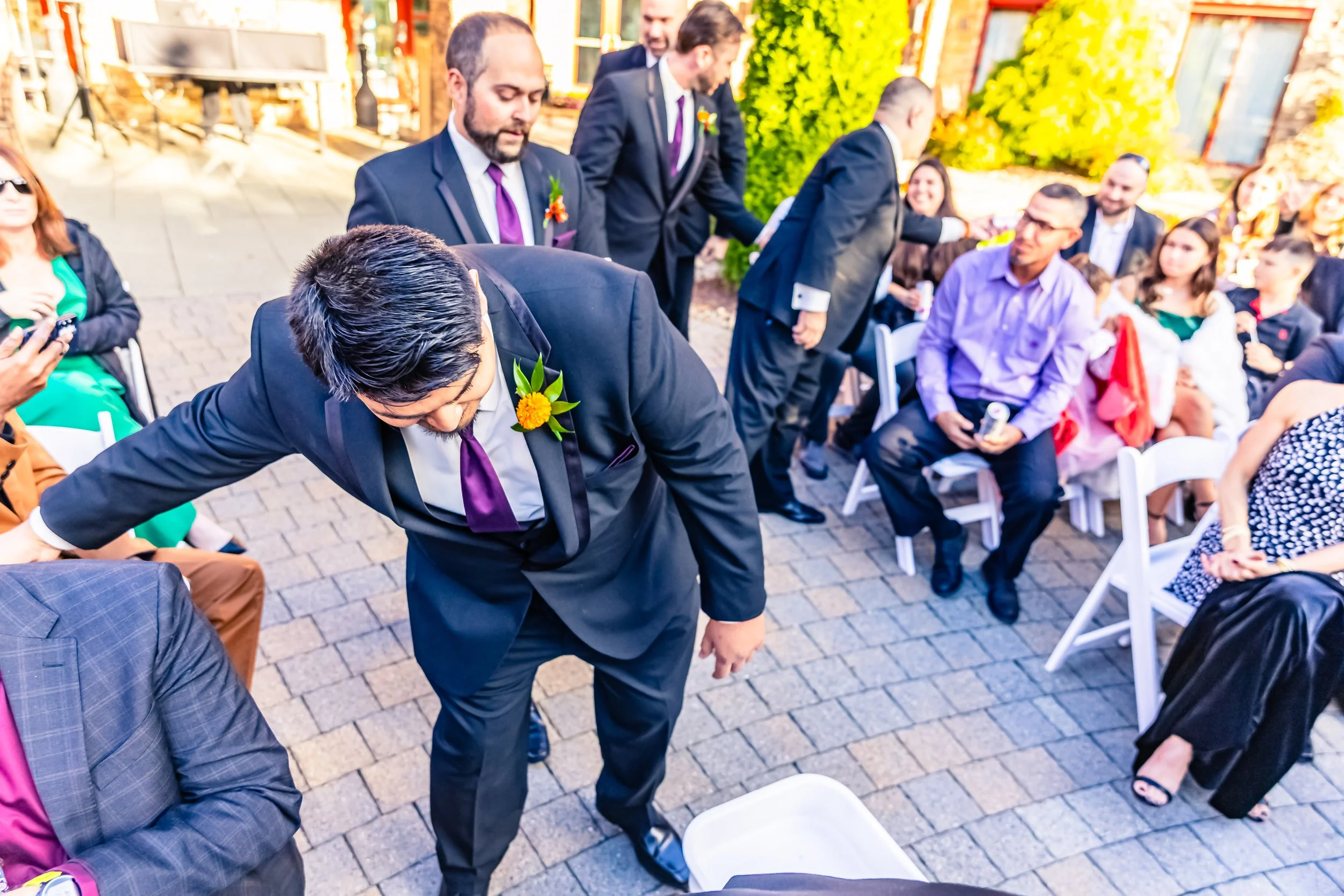 A man in a black suit with a purple tie and a yellow boutonniere leans over to shake hands with a seated guest at an outdoor wedding reception. Other guests are seated or standing around, dressed in formal attire, with some holding drinks. The settin