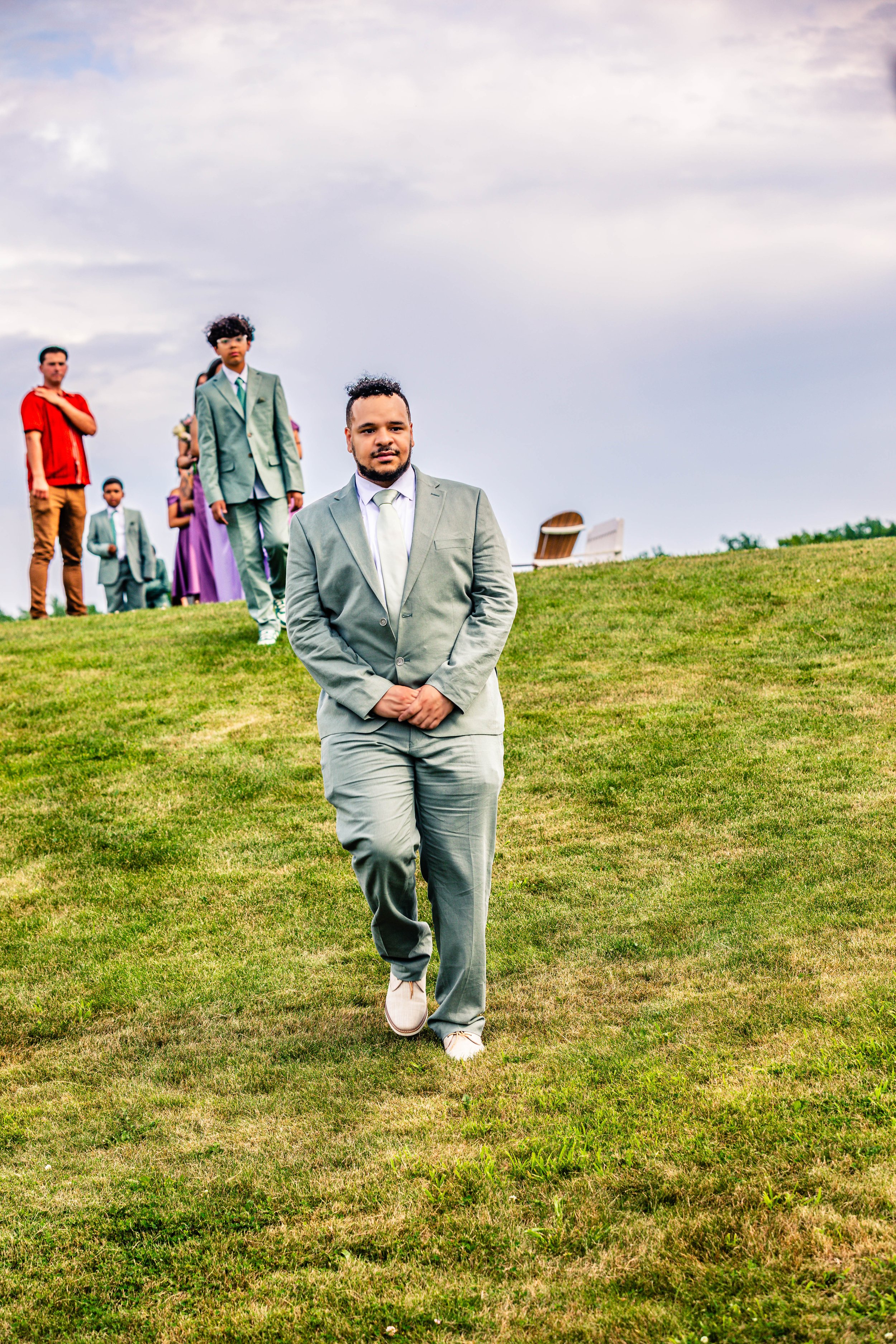 A man in a gray suit walking on a grassy hill, with an outdoor wedding party of six people dressed in colorful and formal attire in the background.