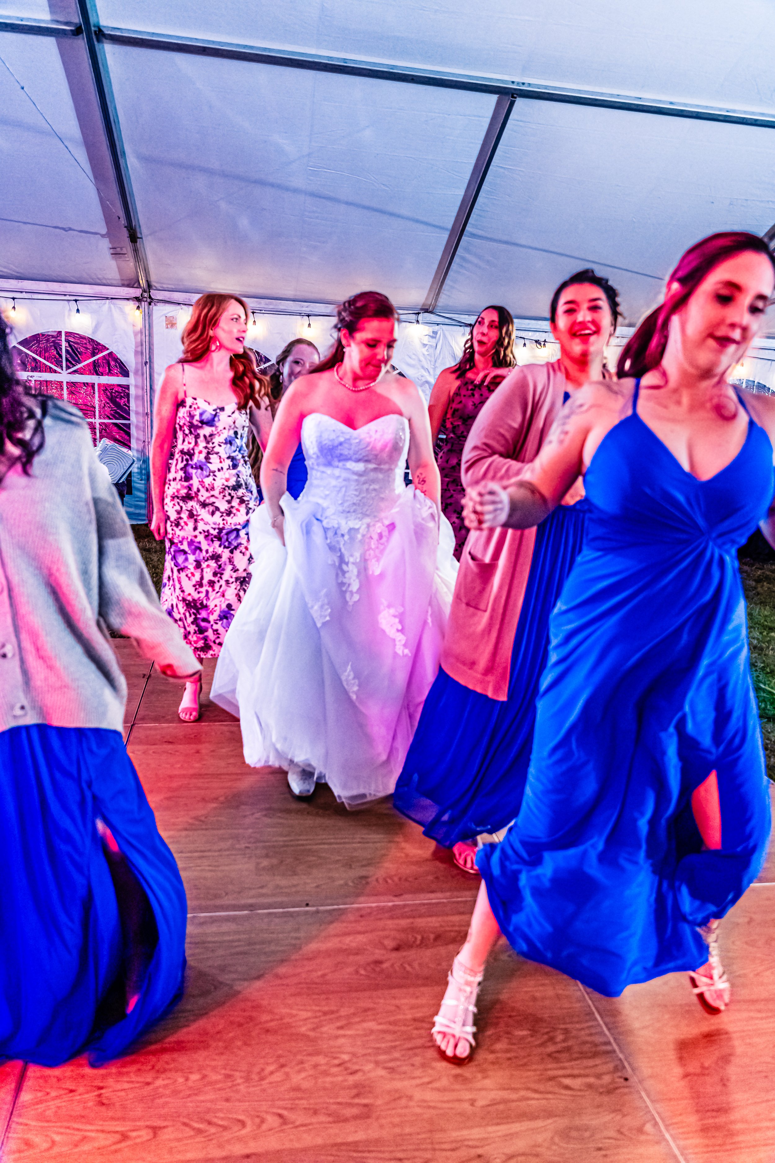 Women dancing inside a tent at a wedding reception, with the bride in a white wedding dress and several bridesmaids in blue dresses.