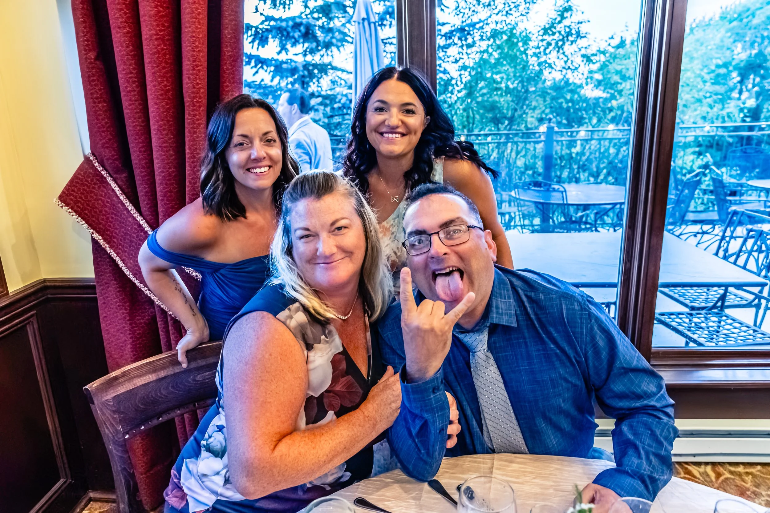 A group of four friends smiling and posing for a photo at a restaurant, with two women standing behind two sitting friends, one of whom is making a playful face and hand gesture.