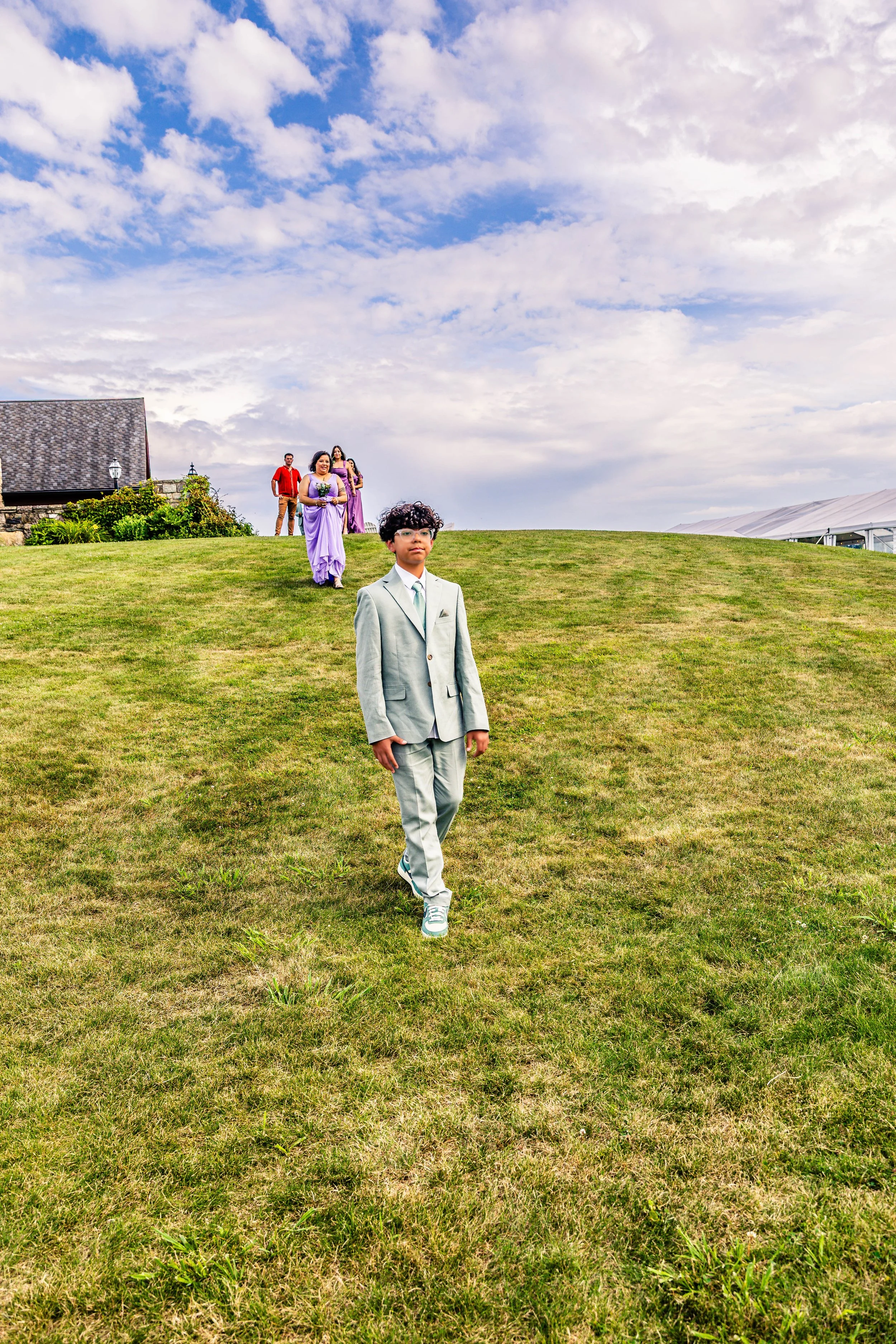A young man in a light gray suit walking on a grassy hill with a group of people behind him, outdoors on a partly cloudy day.