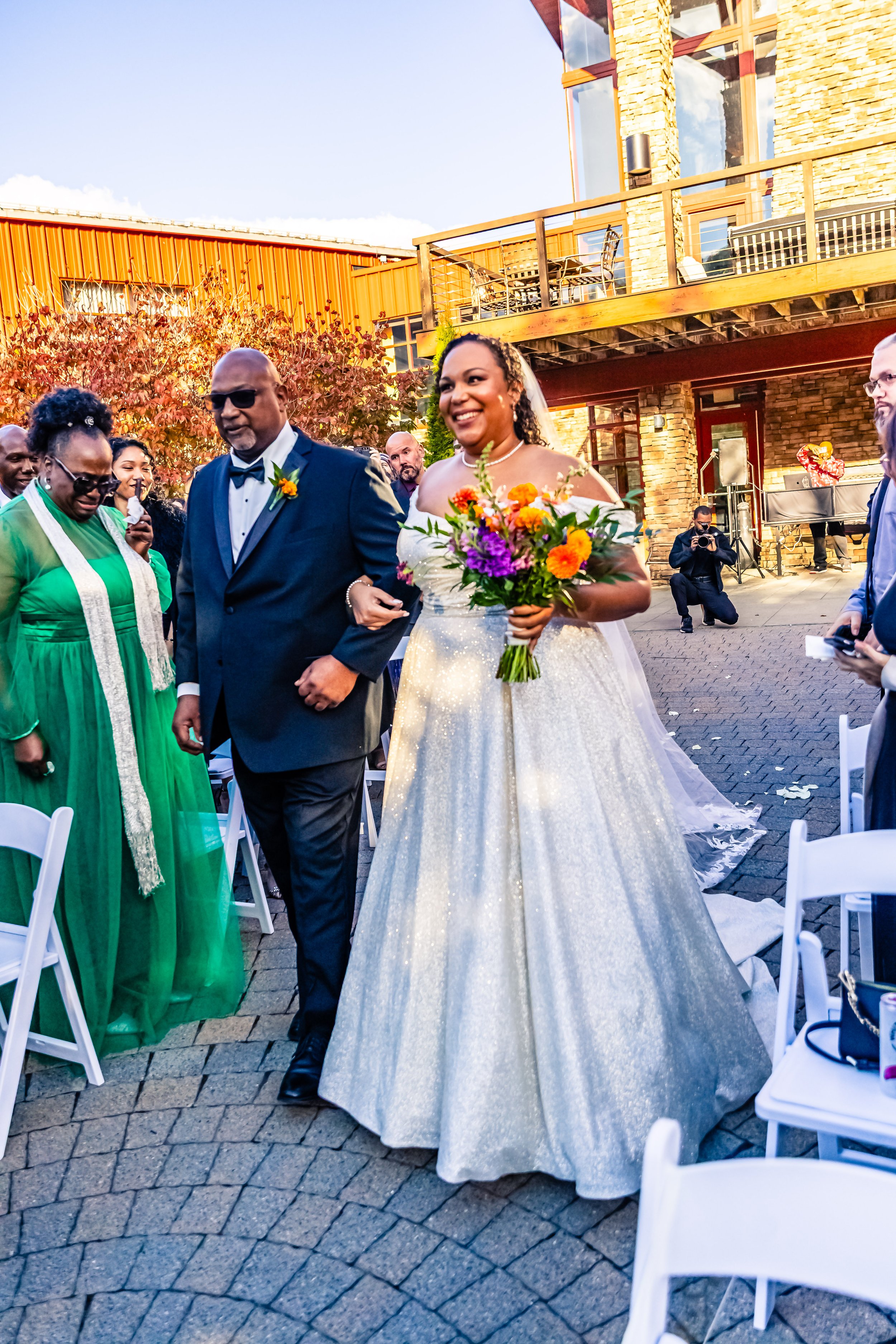 A bride in a white wedding dress holding a colorful bouquet, walking down the aisle with an older man in a black tuxedo, surrounded by wedding guests outdoors.