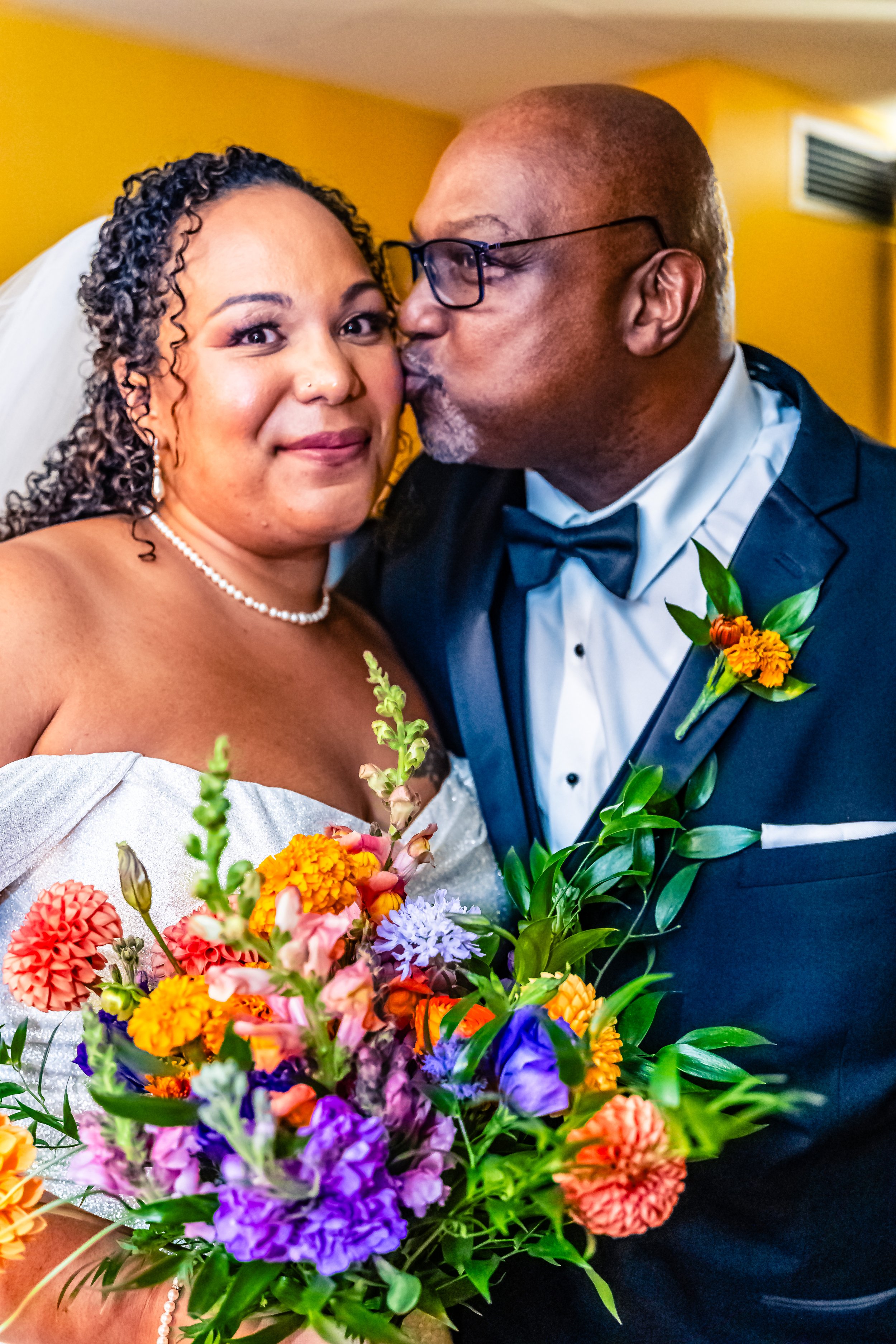 A bride and groom sharing a moment, with the groom kissing the bride's cheek. The bride is holding a colorful bouquet of flowers, and both are dressed in wedding attire.