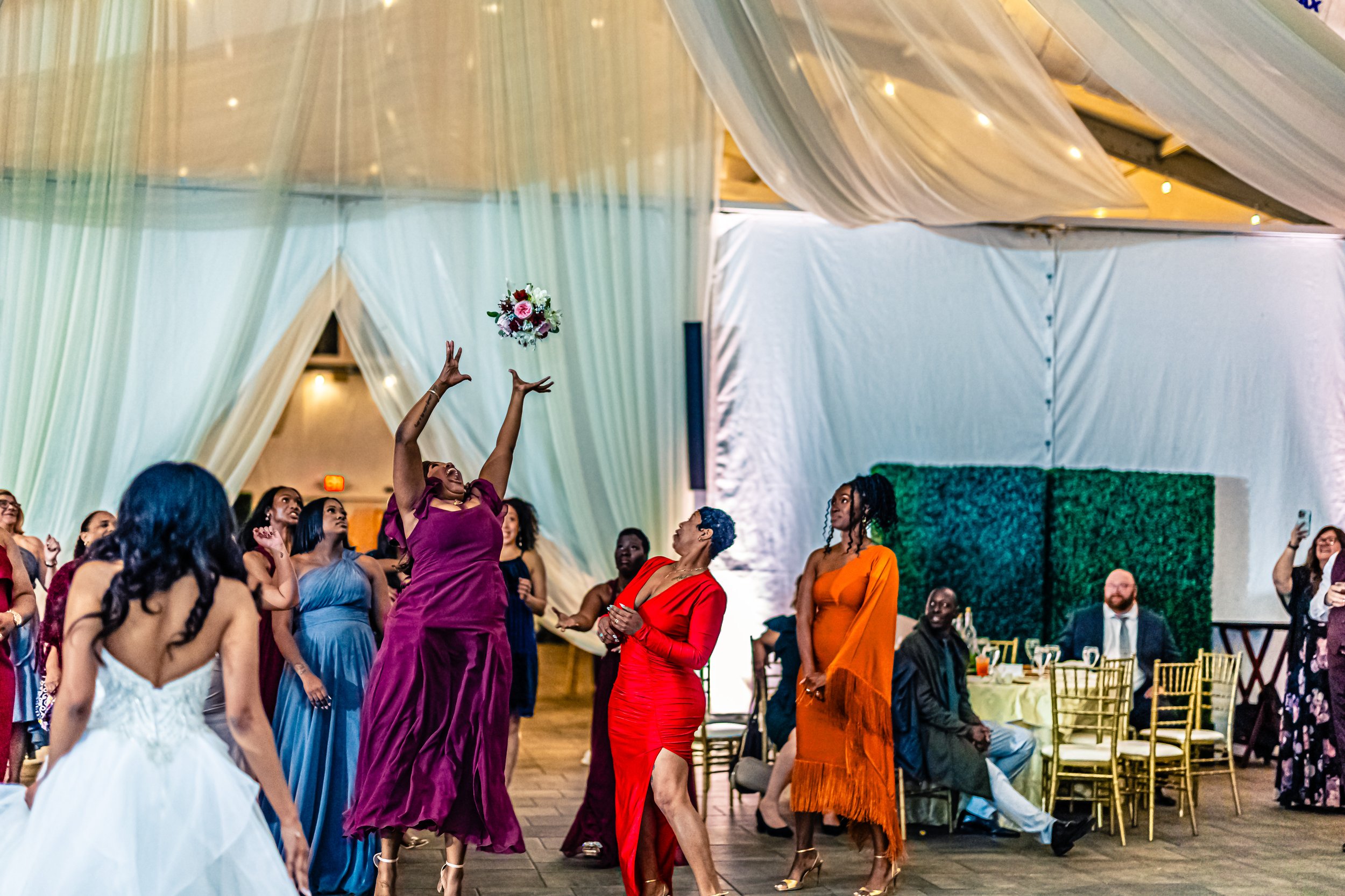 A group of women at a celebration, with one woman in a purple dress tossing a bouquet in the air while others watch and smile; people are seated at tables in the background, at a decorated event venue.