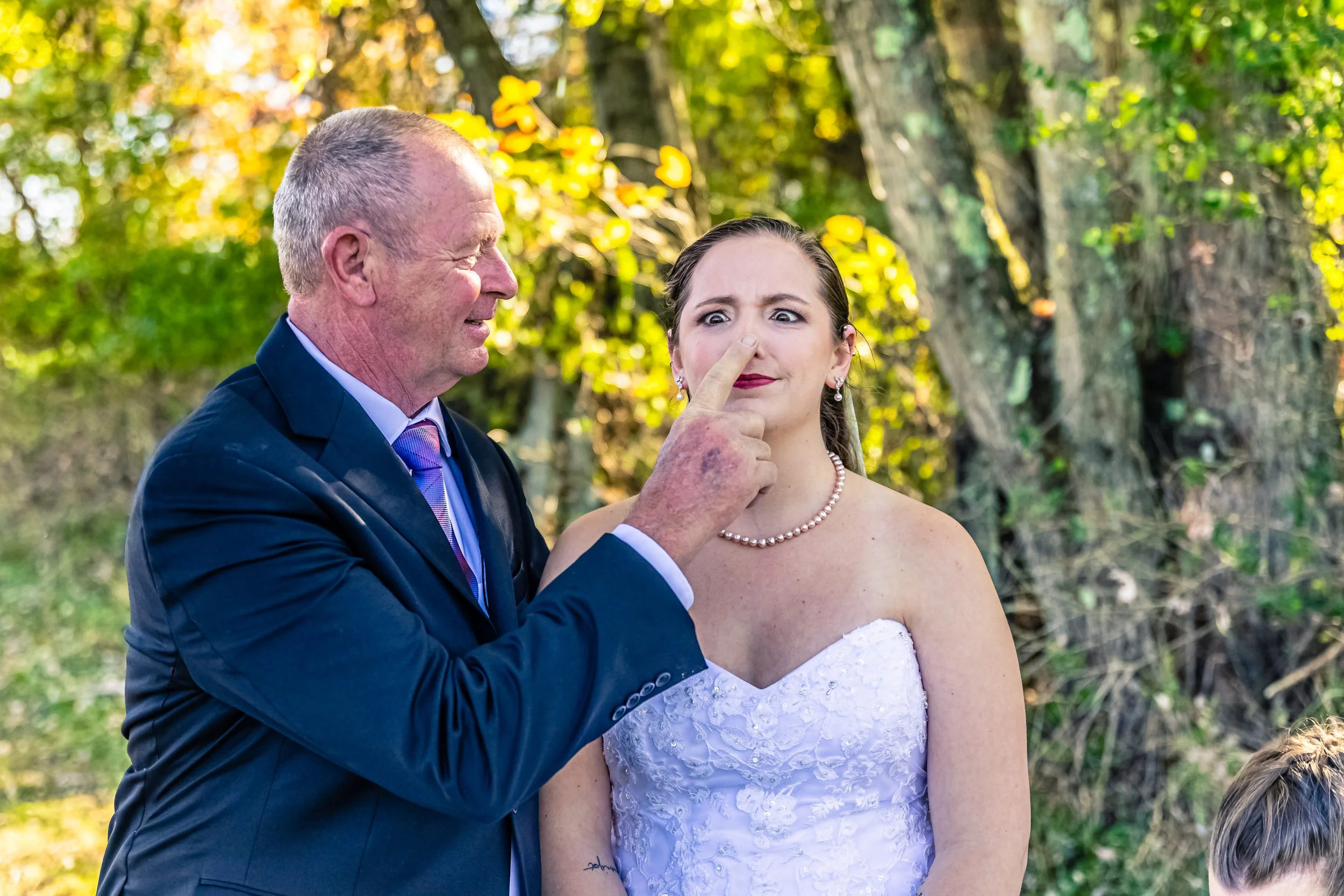 An older man with short gray hair in a dark suit and a woman in a white strapless wedding dress with pearl necklace appears surprised as the man points his finger at his nose, while standing outdoors with green trees in the background.