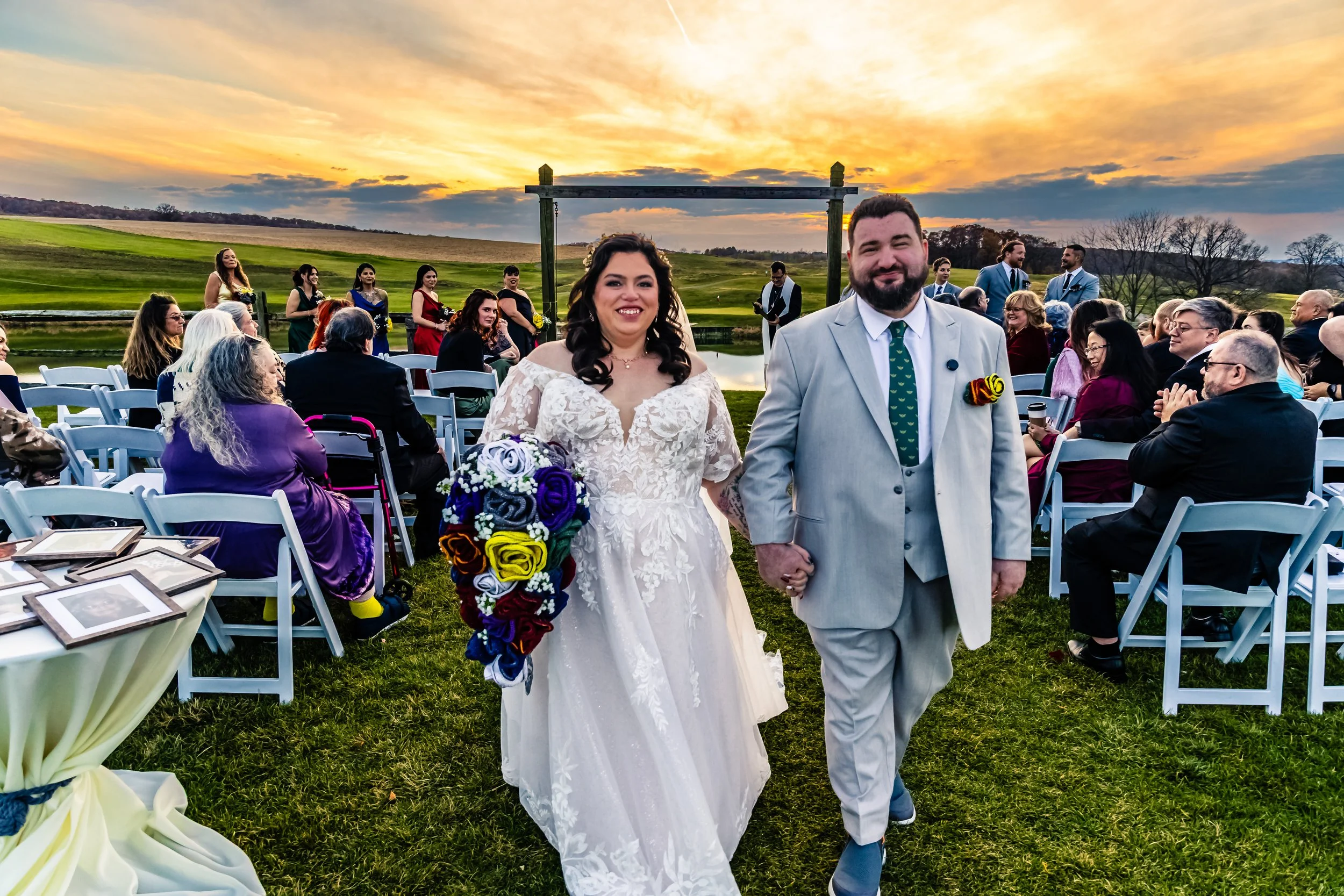 A bride and groom walking hand in hand down the aisle during a sunset wedding ceremony outdoors at a scenic golf course. The bride is wearing a lace wedding gown and holding a colorful bouquet, while the groom is in a light gray suit with a green tie