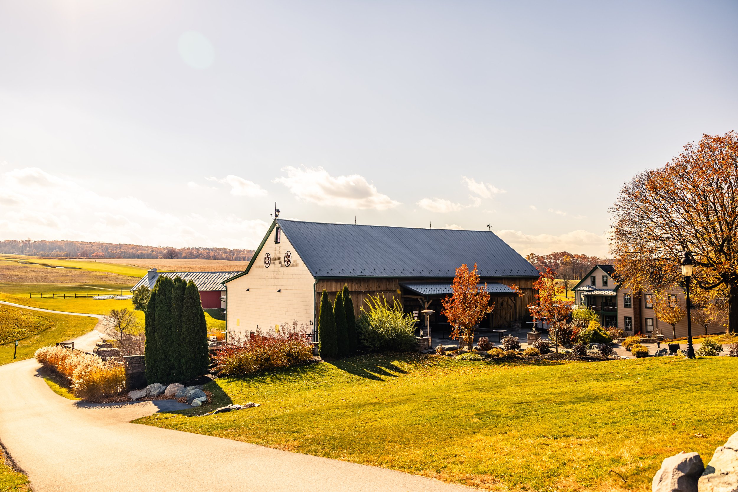 A barn and residential houses in a rural landscape with green fields, trees with fall foliage, and a paved pathway, under a partly cloudy sky.