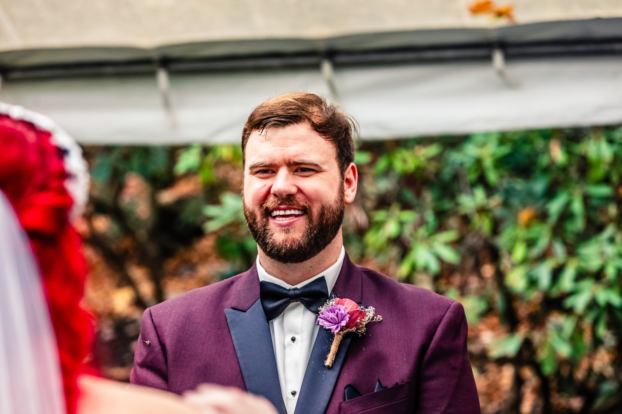 A man with a beard, wearing a tuxedo with a purple jacket, white shirt, black bow tie, and a boutonniere on his lapel, smiling during a wedding ceremony.