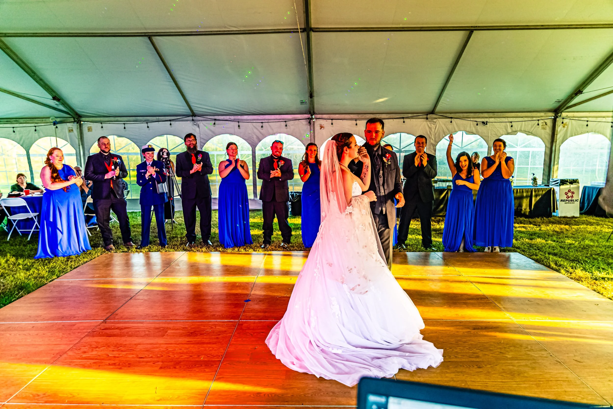 A bride and groom sharing a dance on a wooden dance floor under a large tent, surrounded by bridesmaids and groomsmen applauding and smiling.