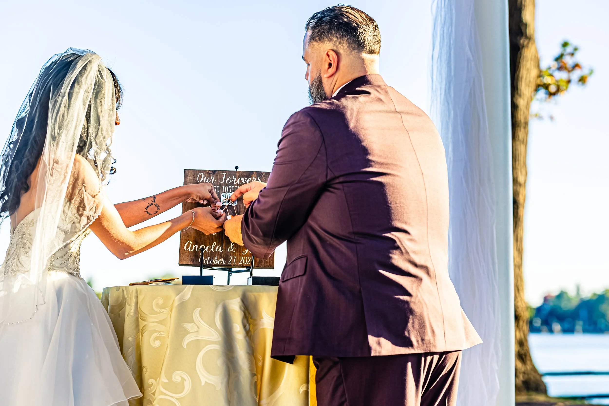 A bride and groom exchanging rings during an outdoor wedding ceremony, with a water and trees in the background.
