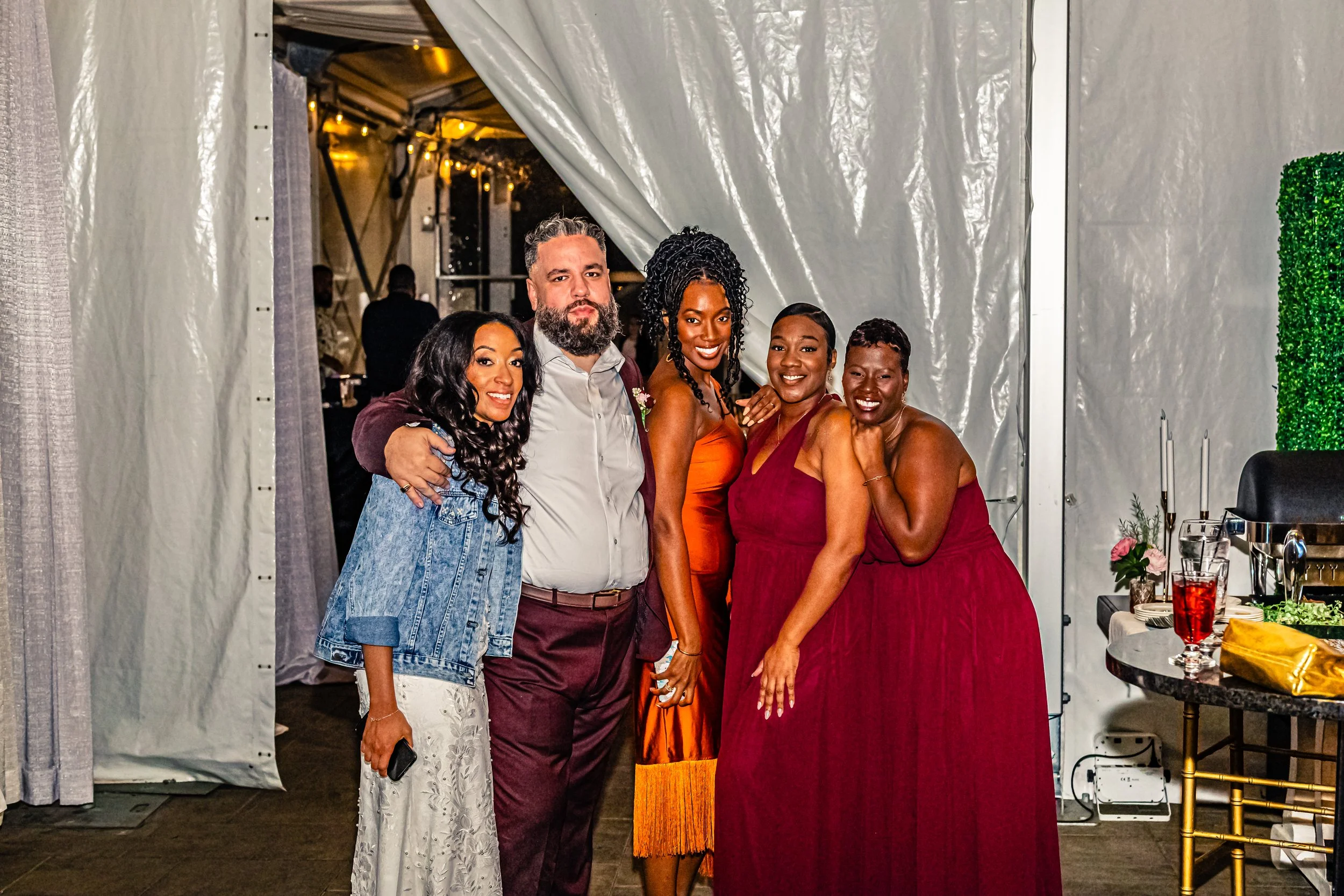 Five people smiling and posing together at an indoor event, with three women and two men dressed in formal and semi-formal attire, standing in front of a white tent wall with warm lighting in the background.
