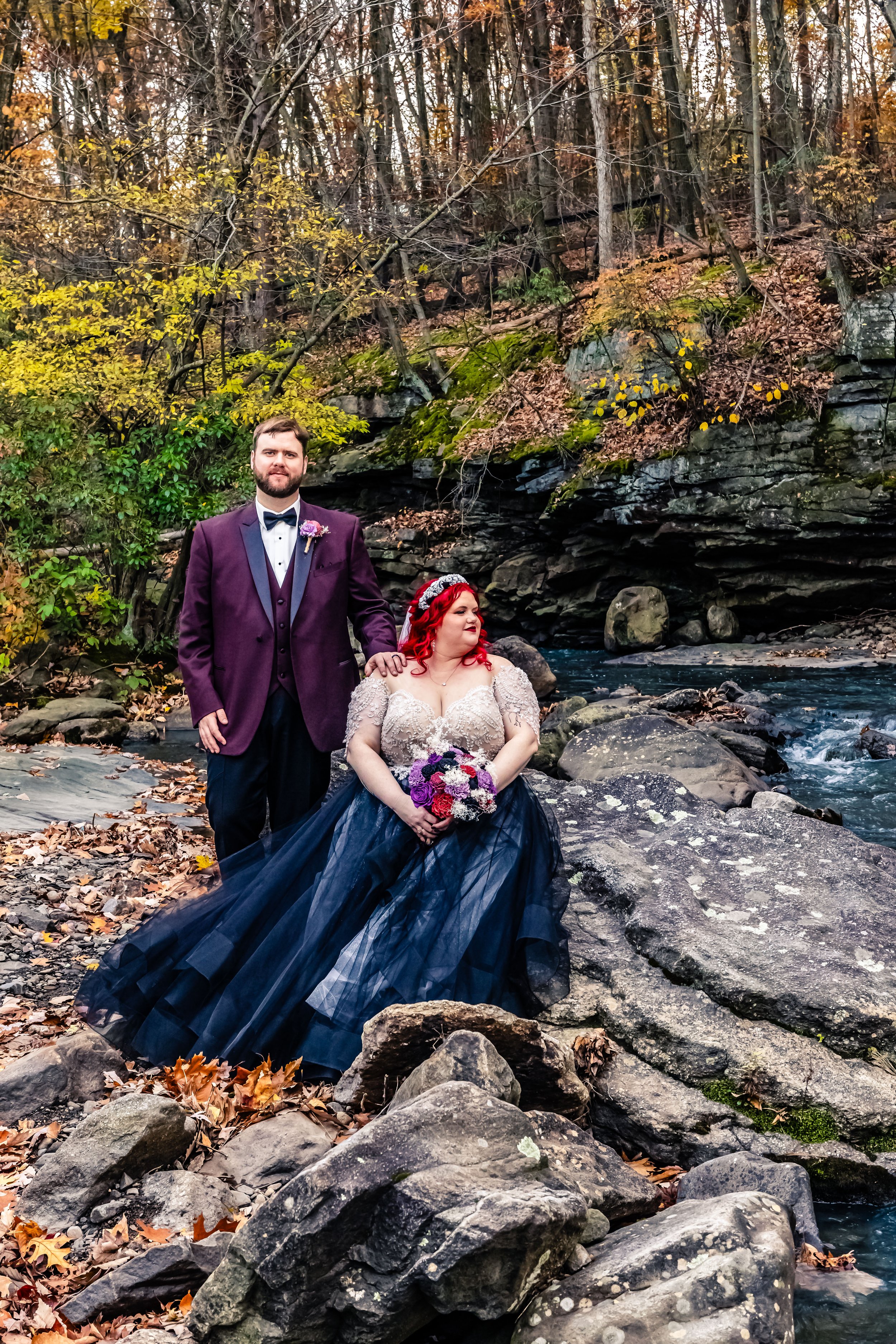 A bride and groom standing by a river in a wooded area during autumn. The bride has red hair and is wearing a dark blue and lace wedding dress, holding a colorful bouquet. The groom has a beard and is dressed in a dark purple suit with a white shirt 