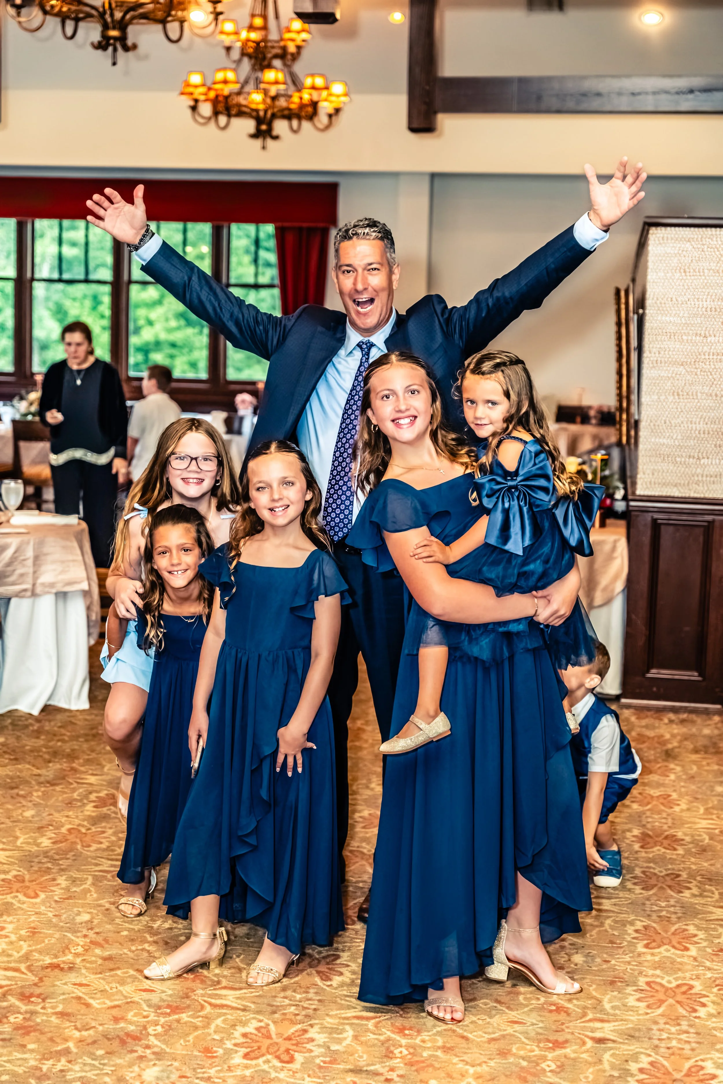 A group of six happy girls in matching blue dresses and an adult man in a blue suit with his arms raised joyfully in an indoor venue, possibly at a celebration or party.
