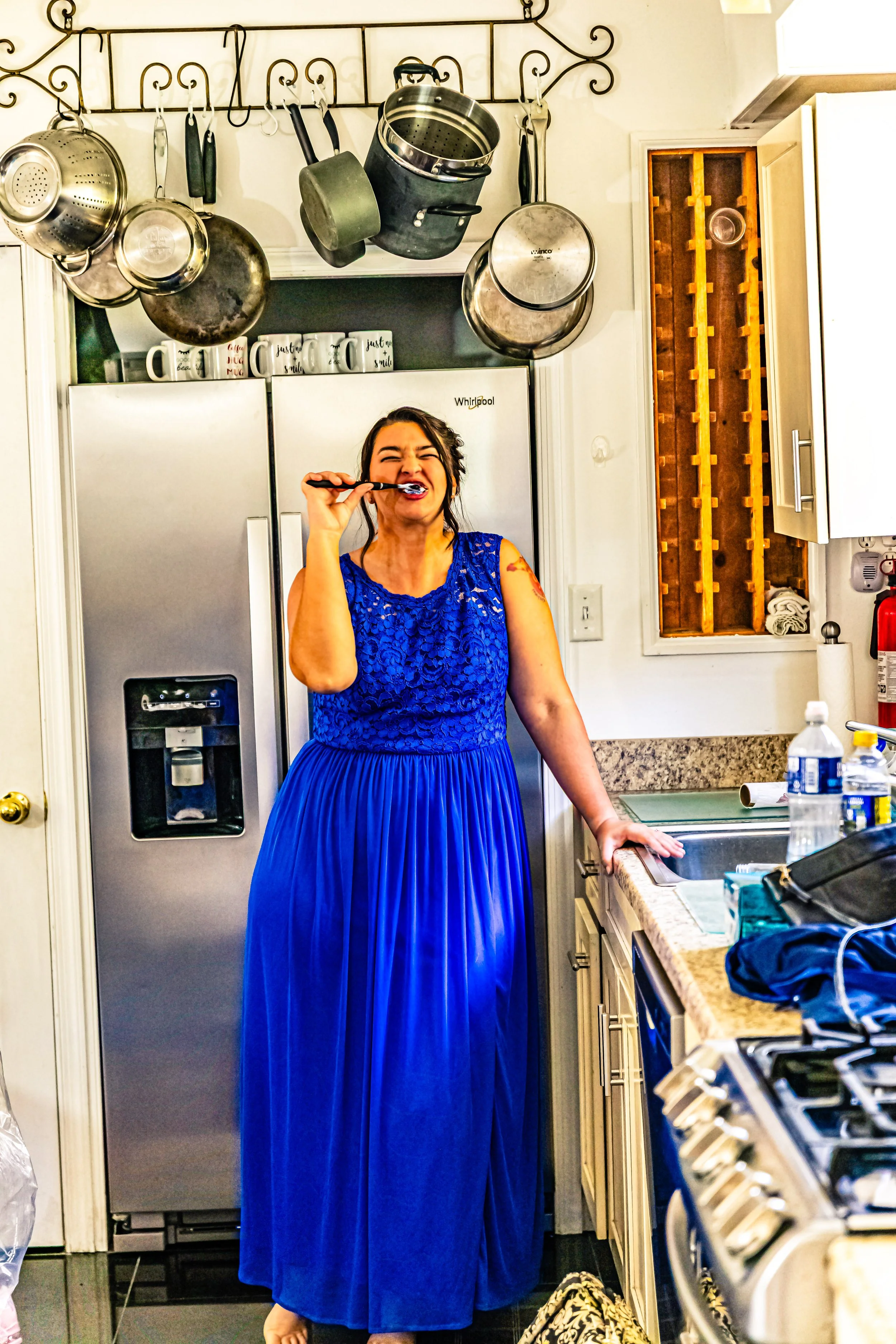 A woman in a bright blue dress brushing her teeth in a kitchen, with pots hanging above her and various household items on the countertop.