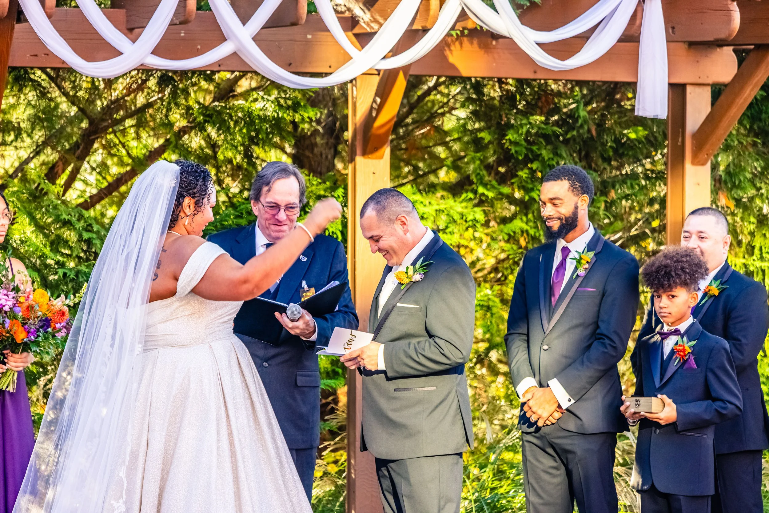 A wedding ceremony outdoors with a bride, groom, officiant, and three groomsmen. The bride is in a white wedding dress and veil. The groom is in a gray suit, holding a waving hand. The officiant and groomsmen are in dark suits with floral boutonniere