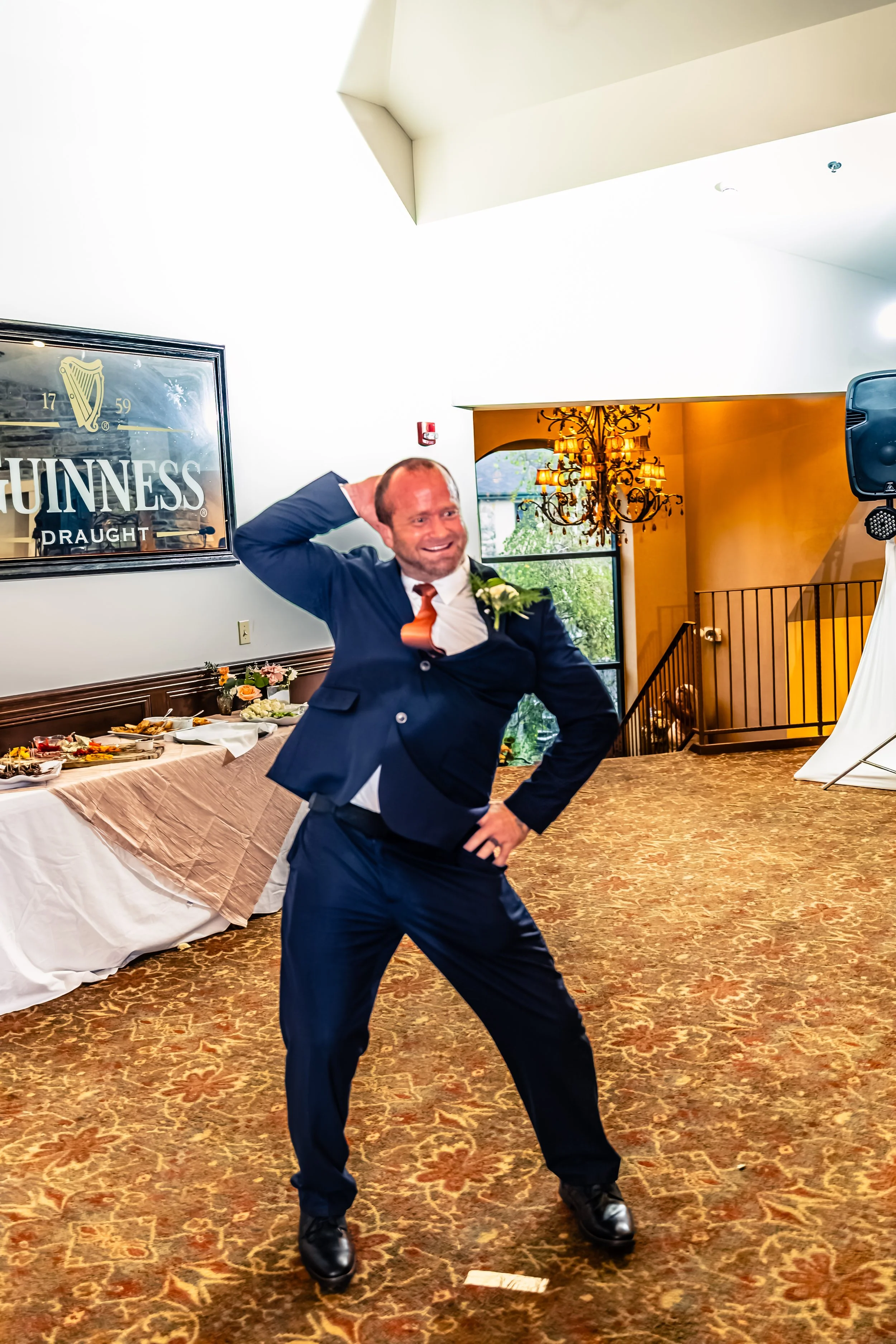 Man in a dark blue suit dancing at a banquet hall with a buffet table and a Guinness sign on the wall.