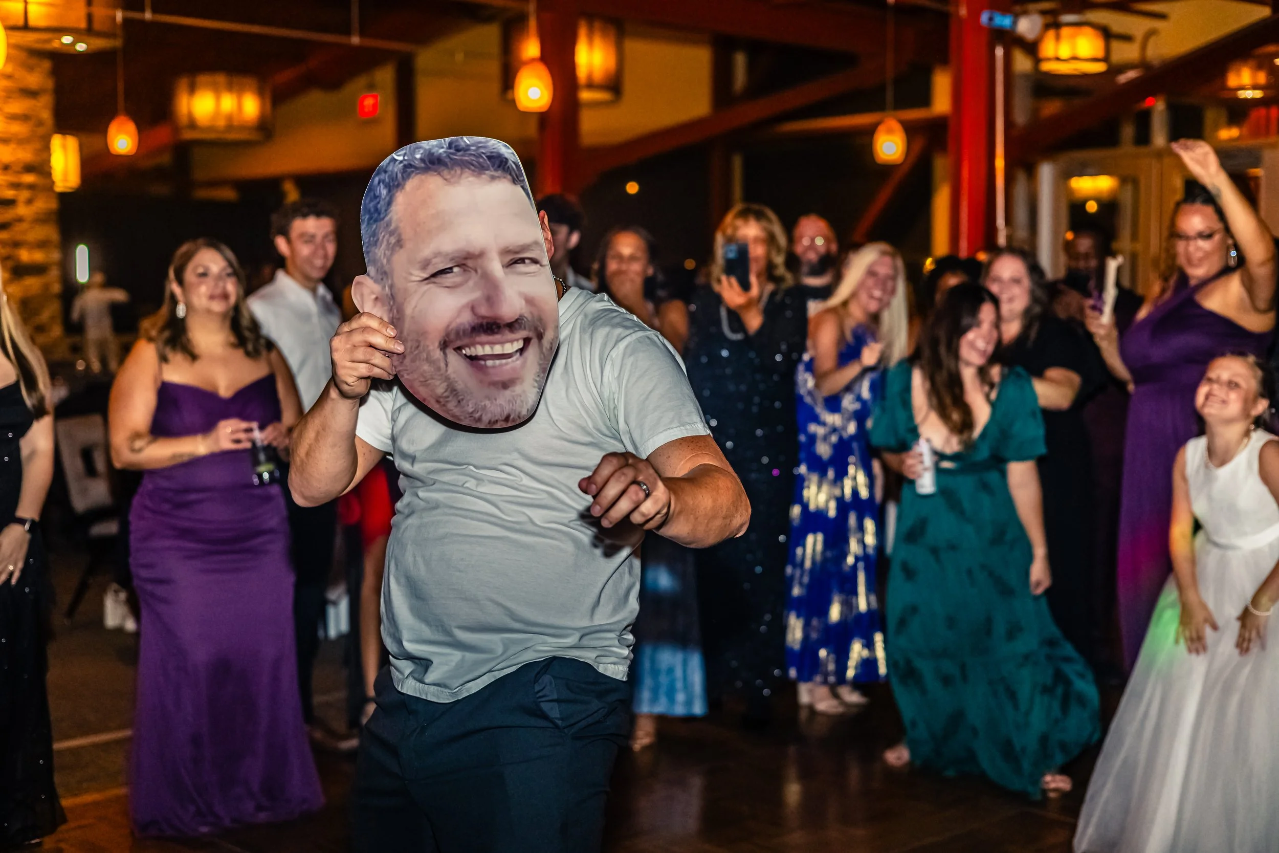 A man wearing a white shirt dancing and holding a large face mask of a smiling man's face at a party with a group of women and children, some taking photos or videos, in an indoor venue with warm lighting.