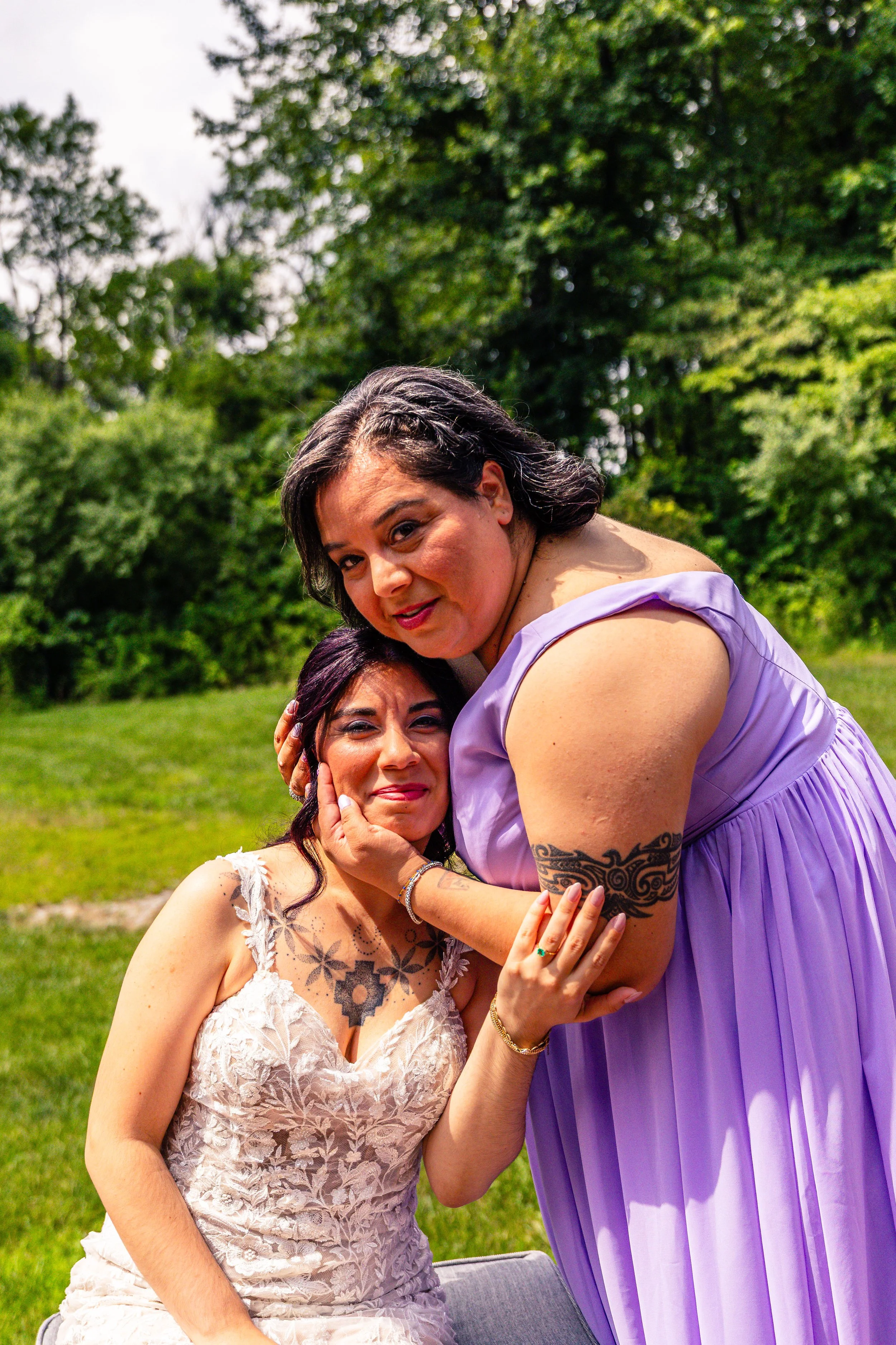 Two women sitting outdoors in a park, one woman with dark hair and a lacy dress, the other woman with dark hair and a purple dress, holding the other woman's face affectionately.