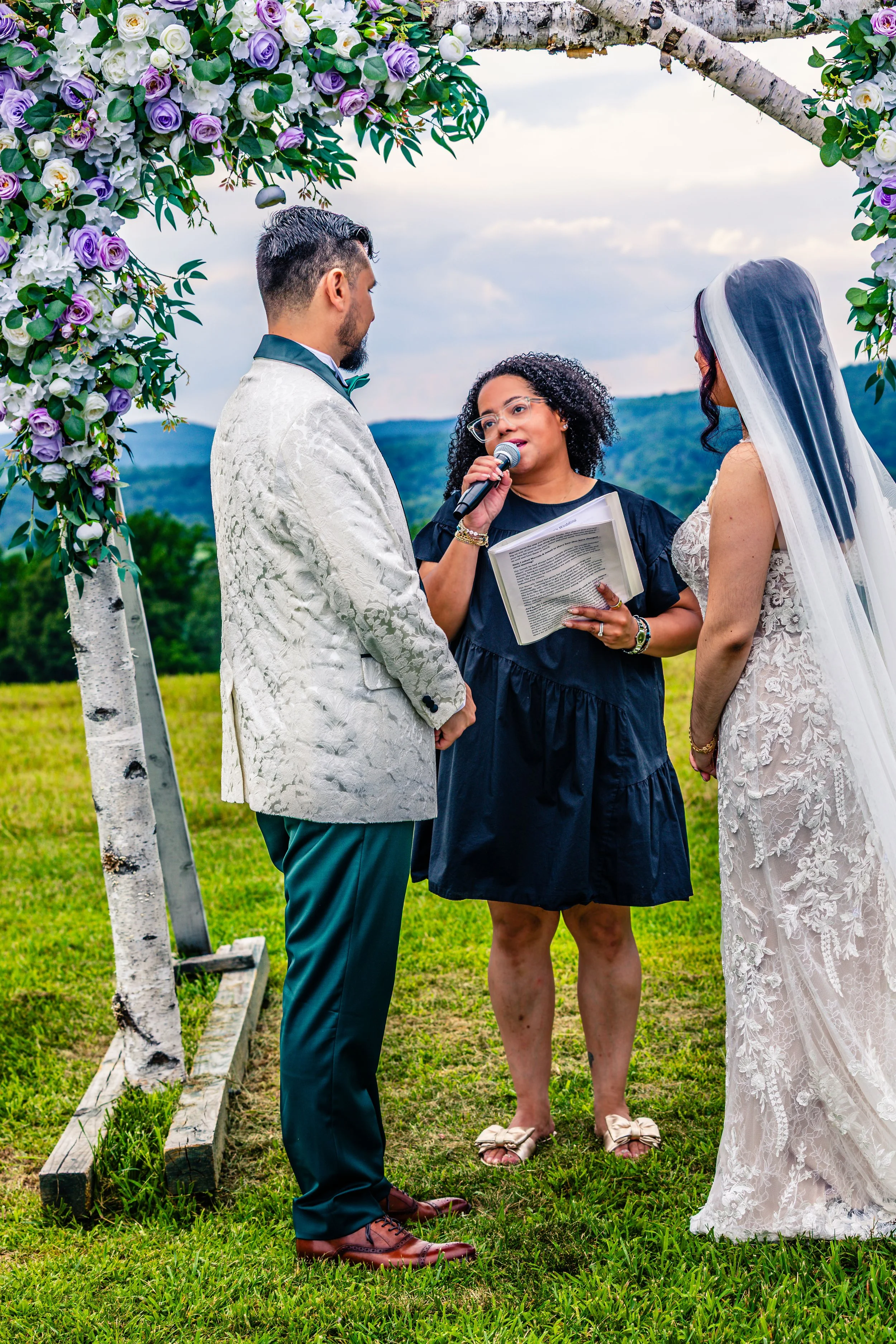 A couple getting married outdoors under an arch adorned with purple and white flowers, with an officiant holding a microphone and a book, in a scenic field with mountains in the background.