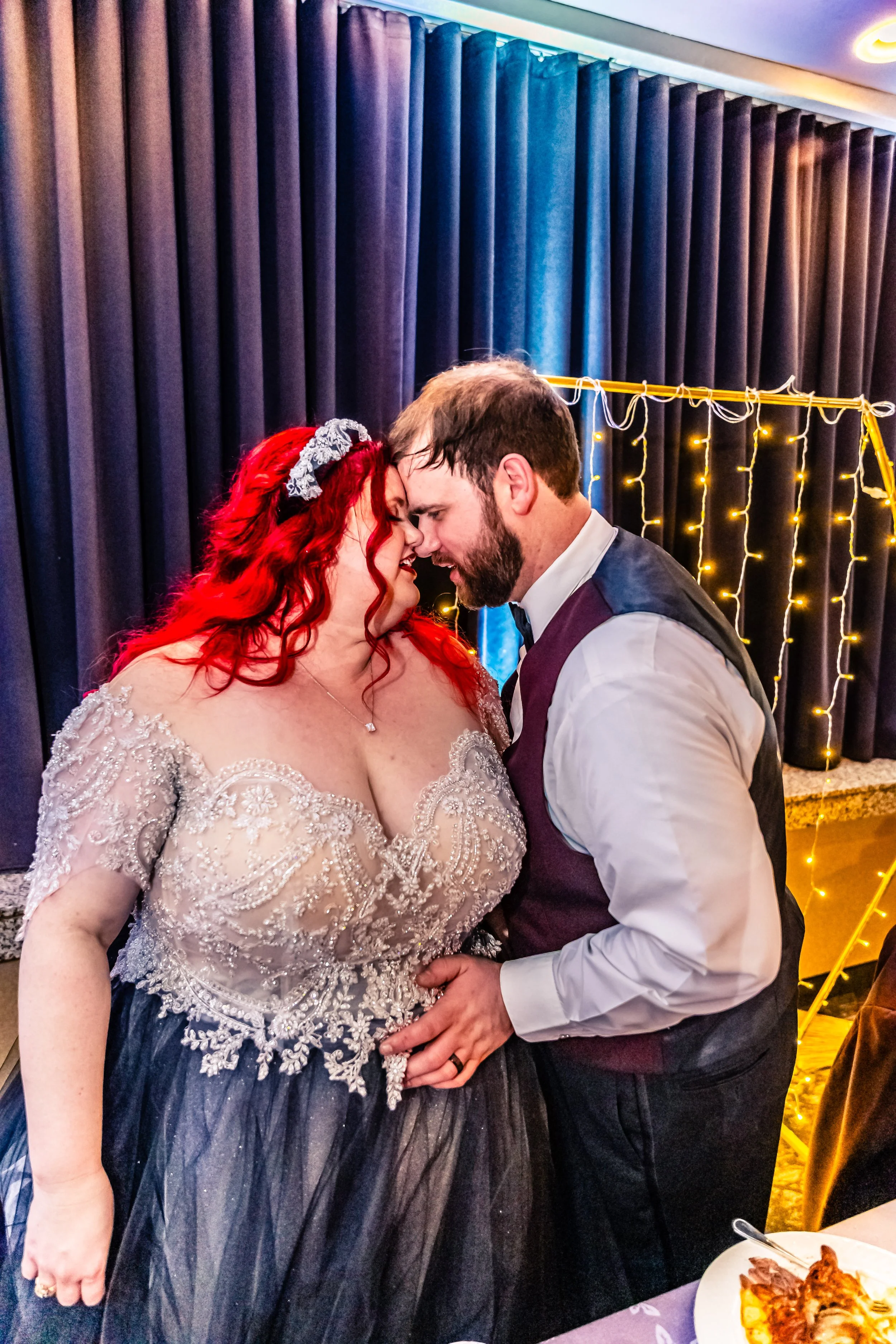 A bride and groom sharing a wedding dance, touching foreheads with eyes closed, in a decorated indoor venue with purple curtains and string lights in the background.