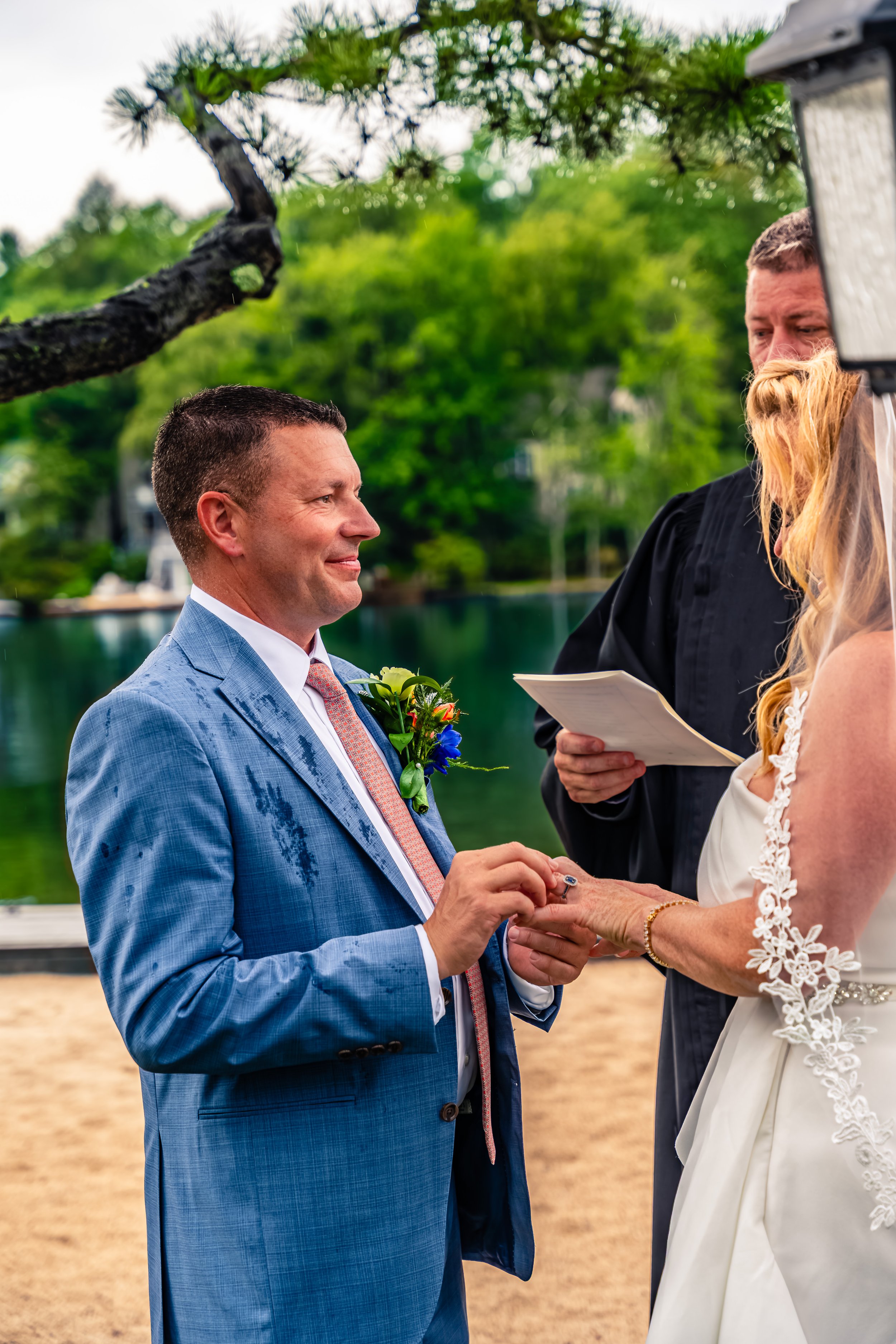 A groom placing the wedding ring on the bride's finger during an outdoor wedding ceremony next to a lake, with an officiant holding a paper and greenery trees in the background.