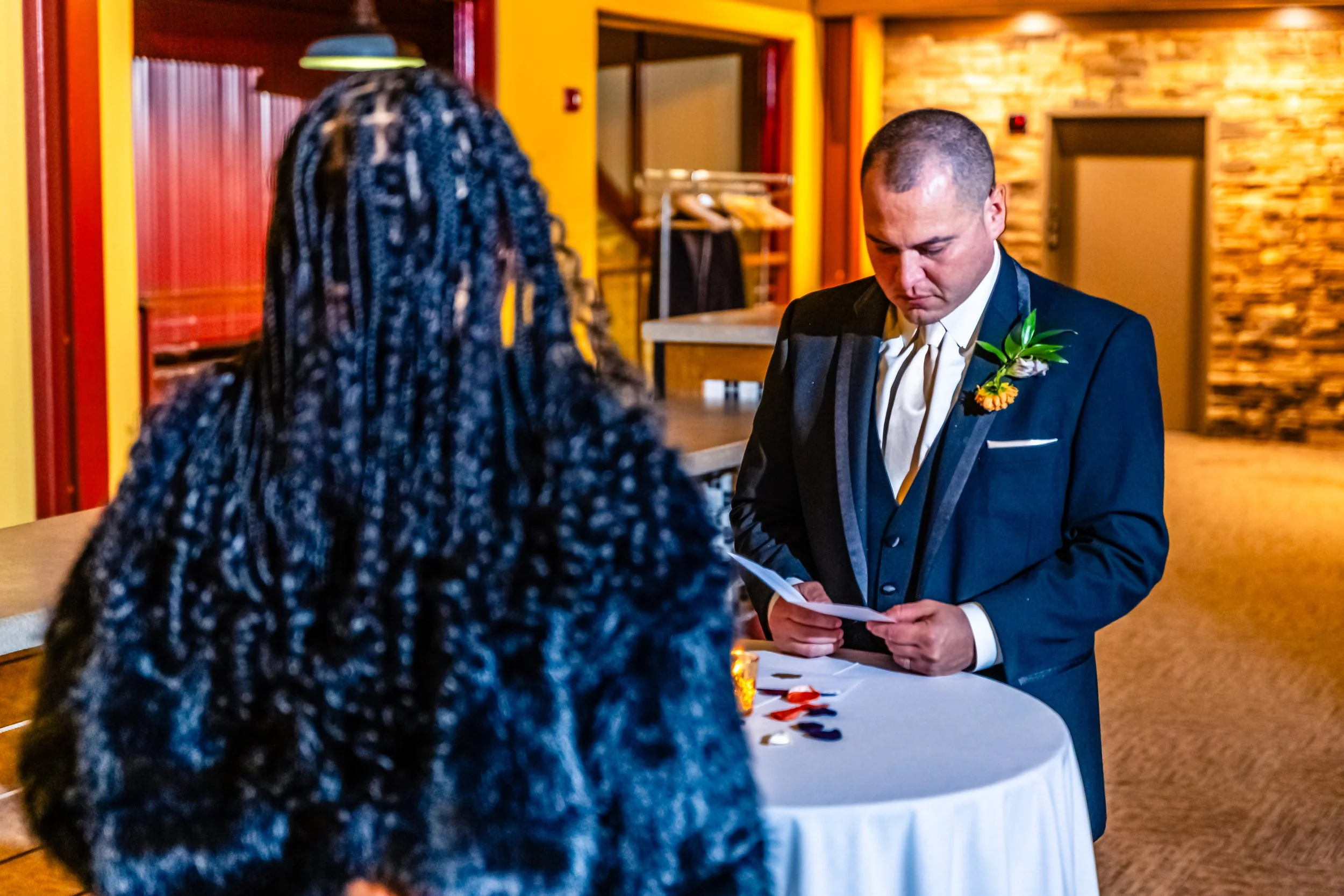 A man dressed in a tuxedo with a boutonniere stands at a white round table, looking down at a piece of paper. A woman with braided hair is standing across from him in an indoor setting with warm lighting and yellow-painted walls.