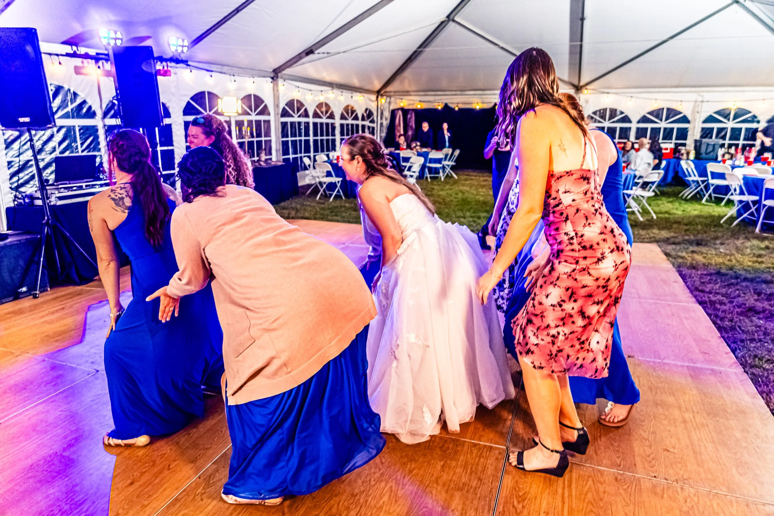 Group of women dancing on a wooden dance floor at an indoor wedding reception, with tables and chairs in the background under a tent.