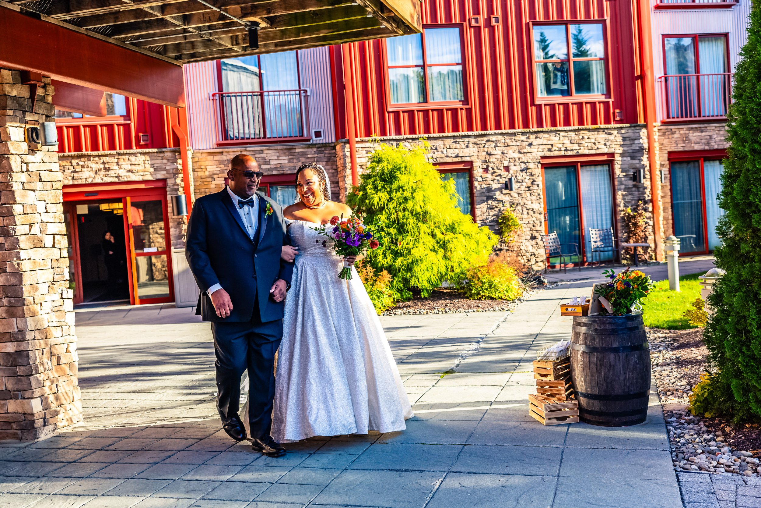 A bride in a white wedding dress walking with a man in a formal suit outside a building with red siding and stone accents, holding a bouquet of colorful flowers, on a sunny day.