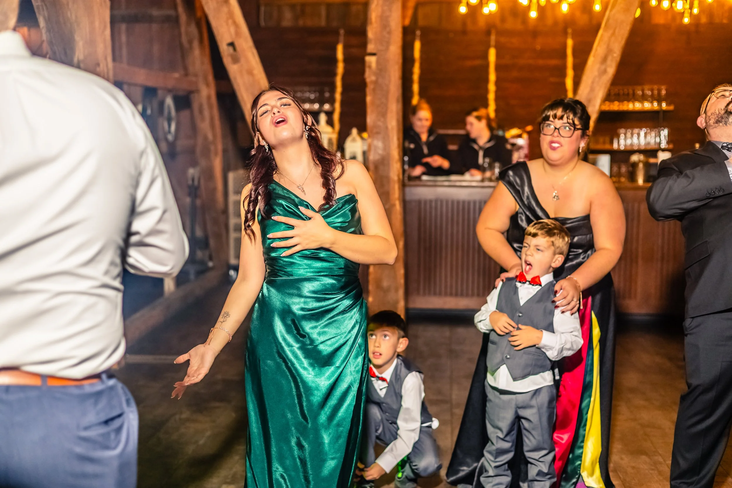 People dancing and enjoying themselves at a wedding reception in a rustic venue with wooden beams and warm lighting, including a woman in a green gown, a woman in a black dress, a young boy in a gray vest and red bow tie, and other guests.