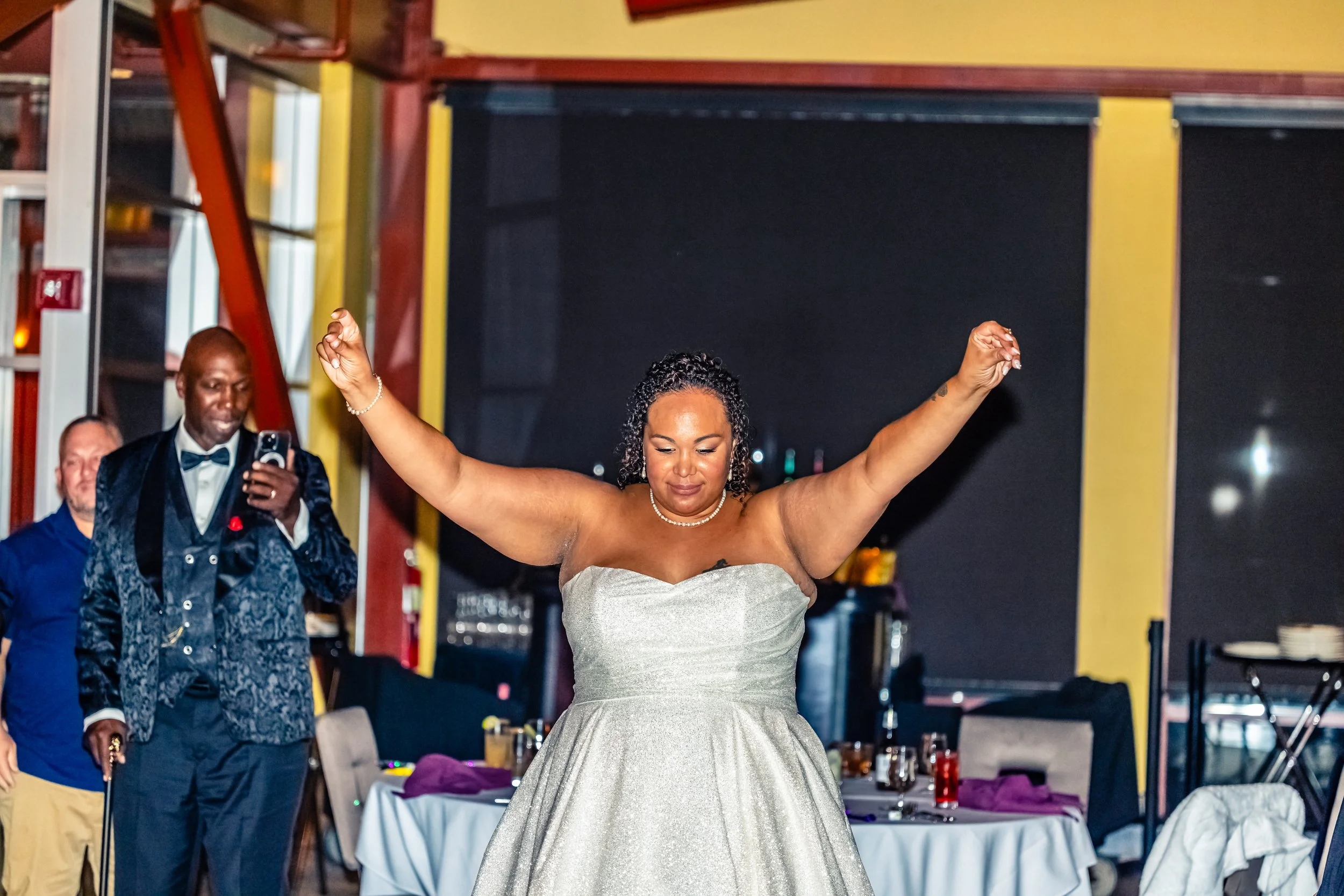 Woman in a strapless silver dress with arms raised, celebrating at a wedding or formal event, with a man in a tuxedo and other guests in the background.