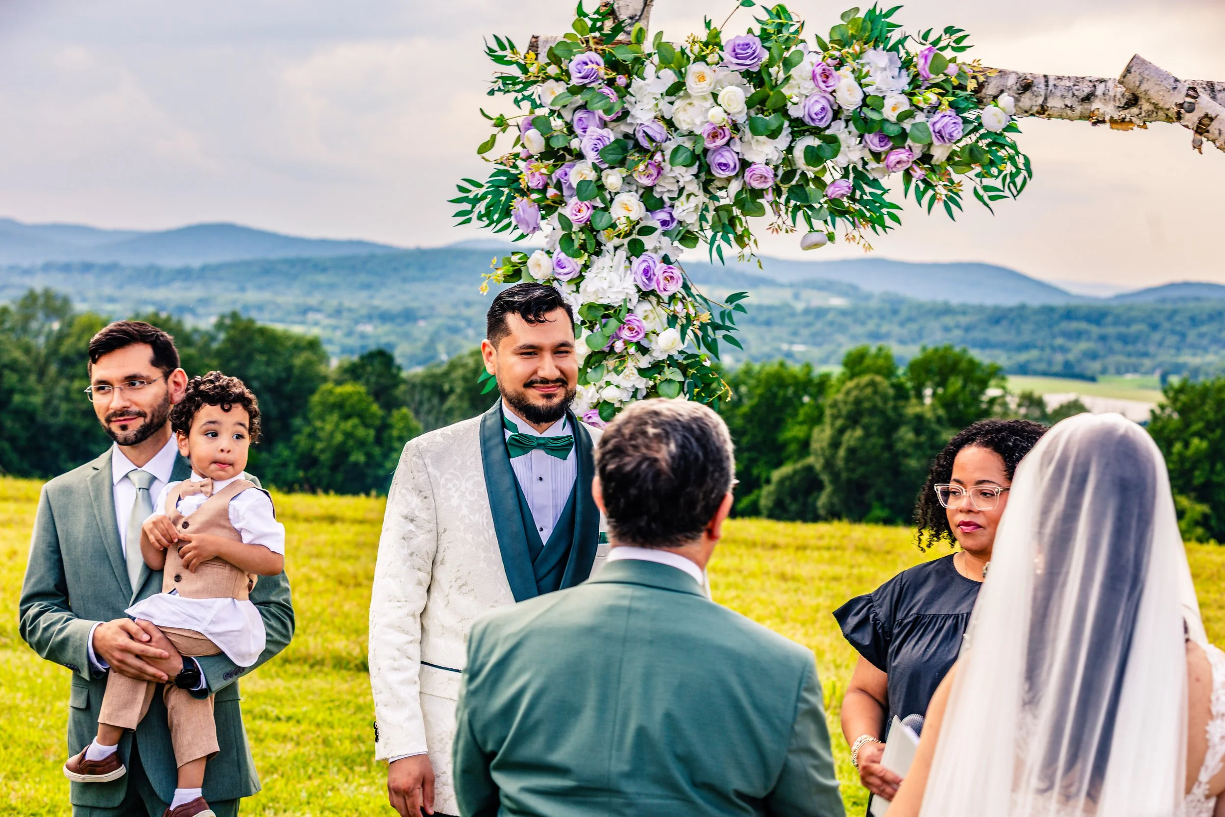 Outdoor wedding ceremony with a groom in a white suit with black lapel and bow tie, standing under a floral arch with purple and white flowers. A bride in a white veil faces the officiant. A woman in black, a man in a green suit, a man with glasses h