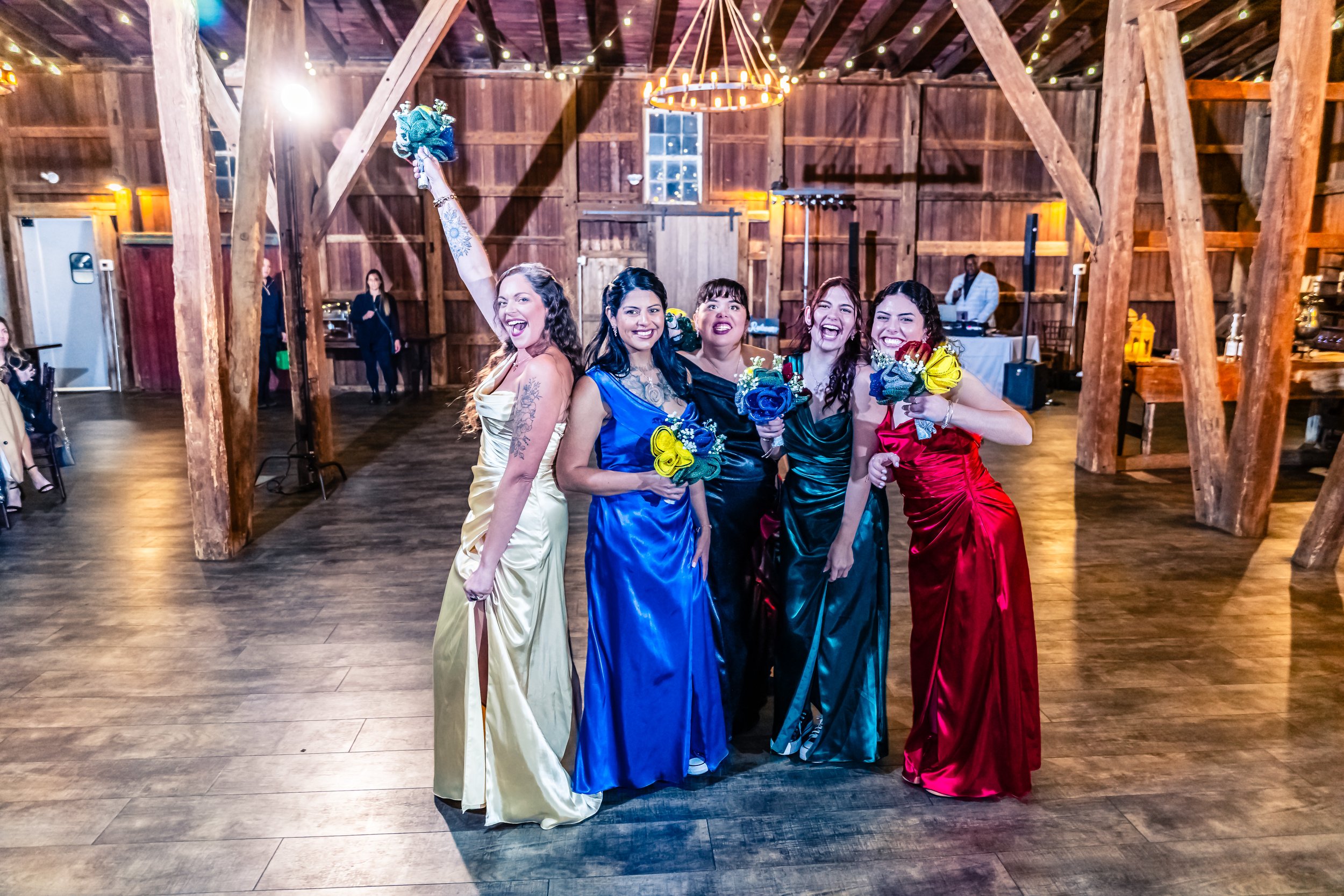 Five women in colorful evening gowns standing together inside a barn-like venue with wooden beams, holding bouquets, celebrating at a wedding or formal event.