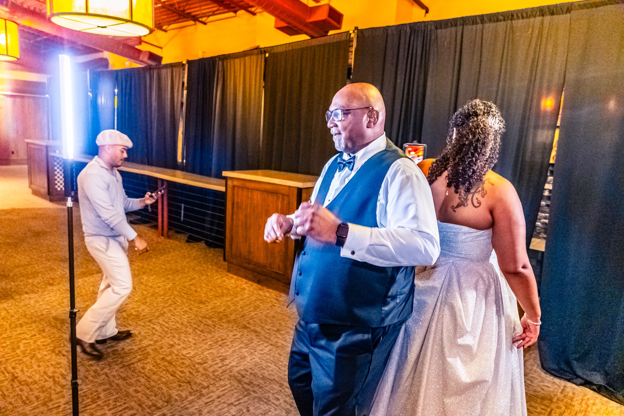 A man in a tuxedo dancing with a woman in a silver dress at a party, while a person in white with a beret looks at their phone in the background.
