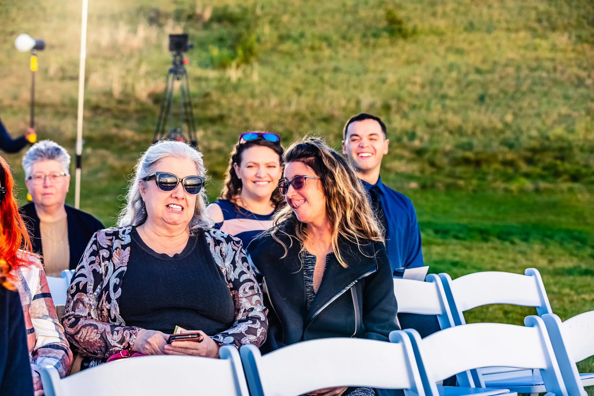Group of people sitting outdoors, laughing and smiling at an event, with chairs and a grassy hill in the background.