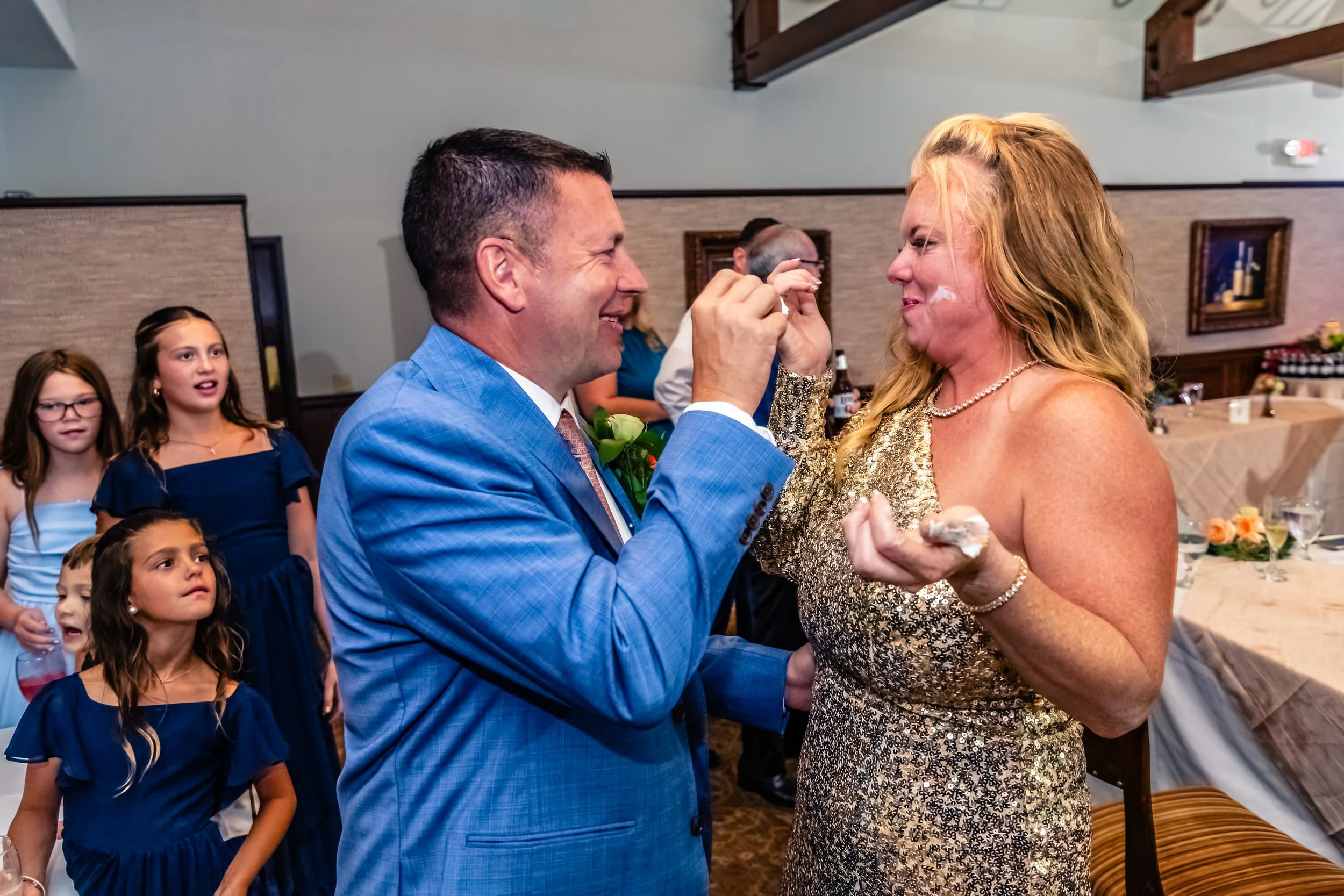 Man and woman at a celebration, the man is feeding the woman cake, surrounded by children at a party or wedding.