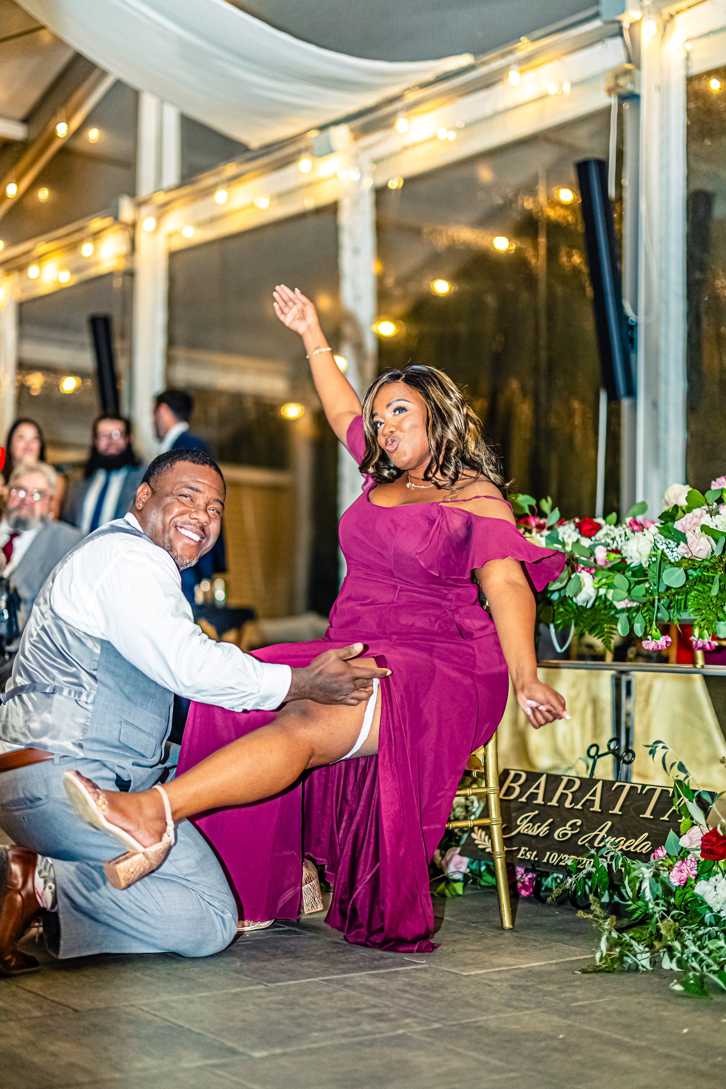 A man lifting a woman at a wedding reception, both smiling, with guests and decorations in the background.