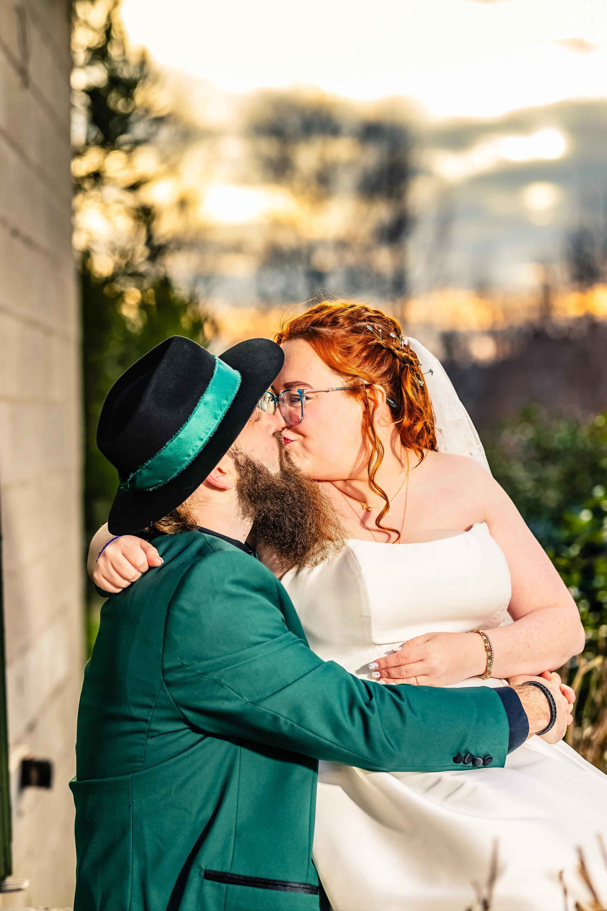 A couple at their wedding, kissing while embracing outdoors during sunset.